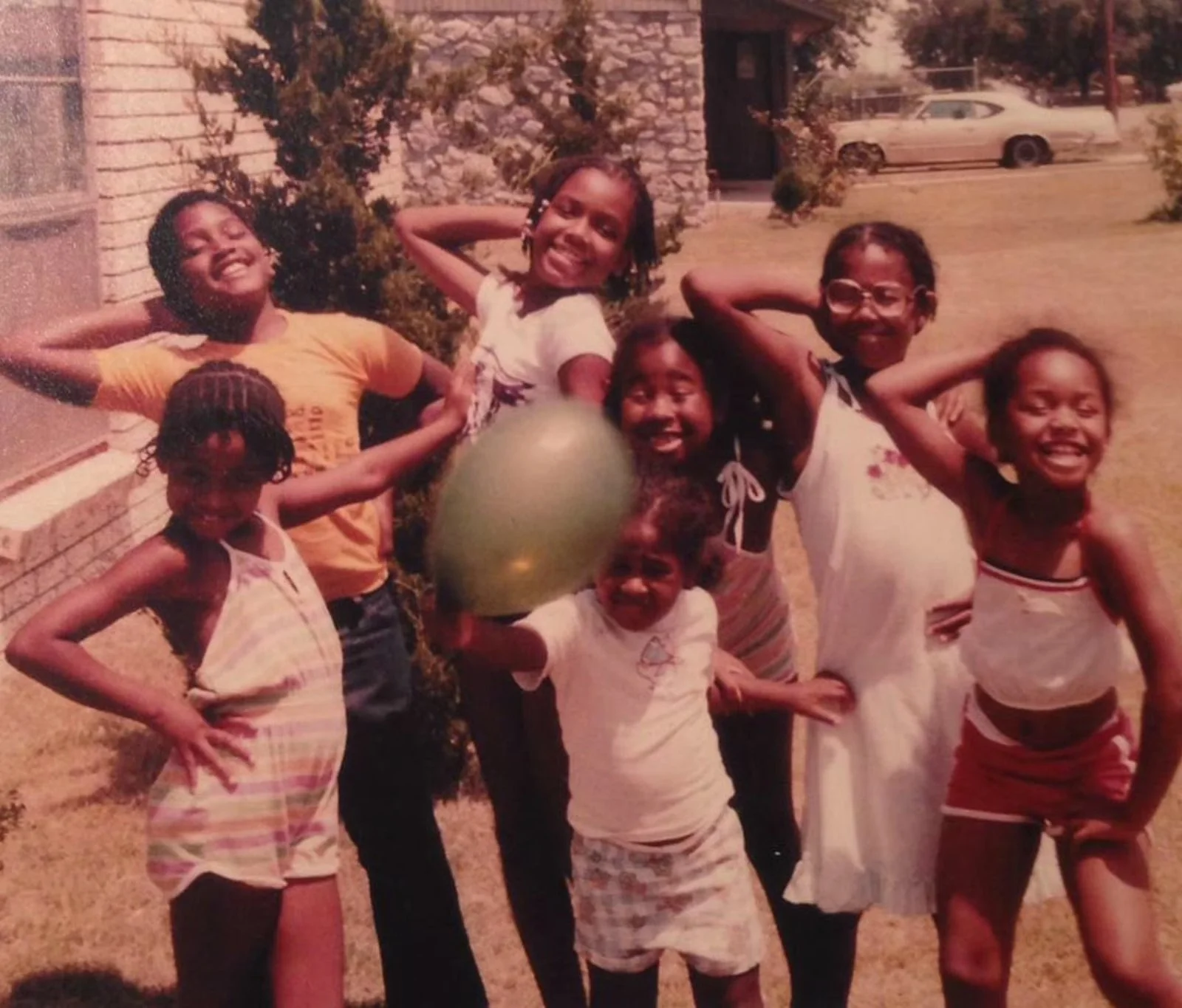 Group of six children smiling and posing outdoors, some with hands on hips, one holding a green ball, in front of a brick building, trees, and a parked white car.