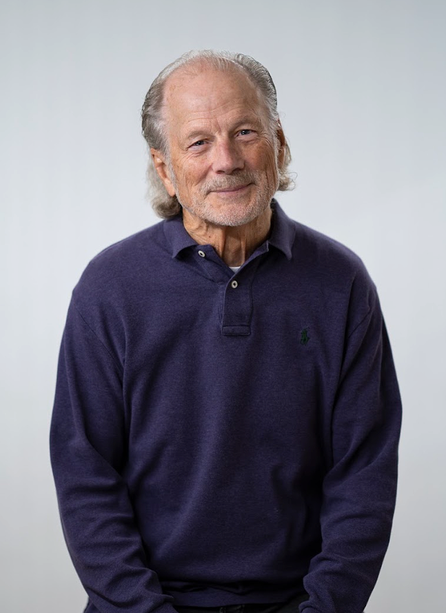 An elderly man with gray hair and a beard wearing a dark blue collared shirt, standing against a plain light gray background.