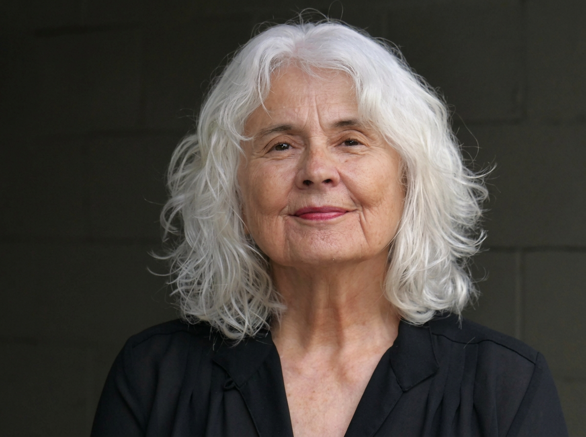 A smiling elderly woman with curly white hair and fair skin, wearing a black top, standing against a dark gray background.