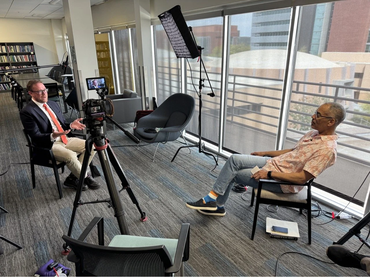 Two men are having an interview in a studio. One man, dressed in a blazer, shirt, and khakis, is speaking while gesturing with his hands. The other man, wearing a patterned shirt, jeans, and colorful socks, is sitting with a notepad and a book on his