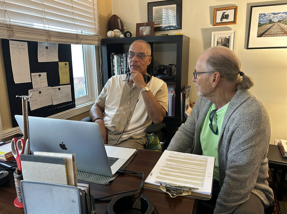 Two men sit at a desk in a home office, engaged in a conversation. One man is wearing a beige button-down shirt, glasses, and headphones around his neck, thinking with his hand on his chin. The other man is wearing a gray sweater, glasses, and has long hair tied back, speaking to him. The desk has a silver MacBook, legal pads, a clipboard, and office supplies. Behind them are framed pictures, bookcases, and a window with blinds.