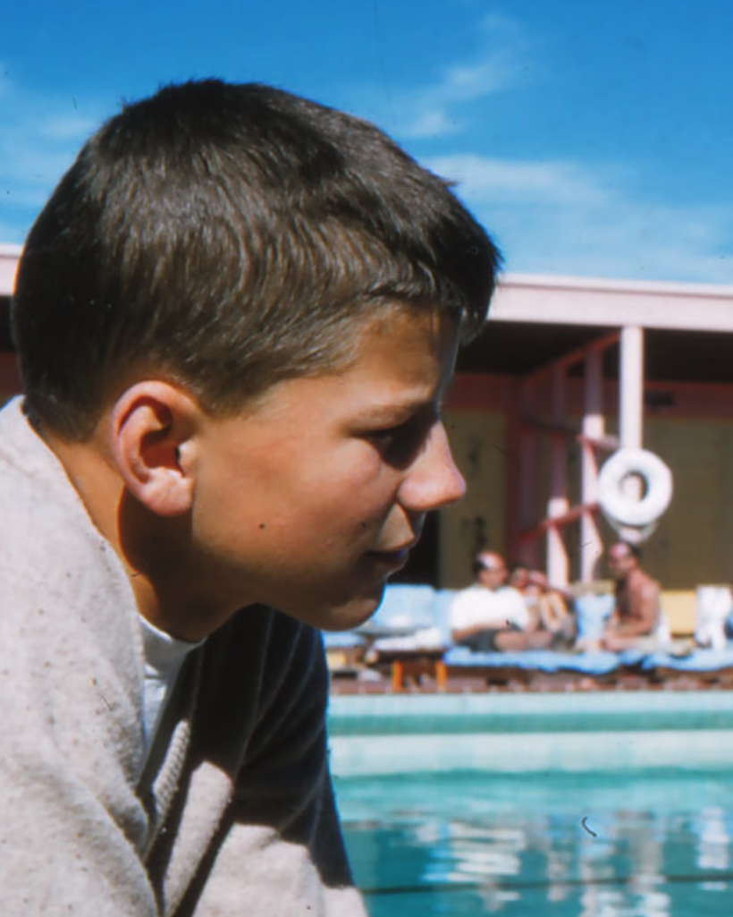 Close-up side profile of a young man with short dark hair, wearing a gray hoodie, at a poolside with people lounging in the background, under a blue sky.