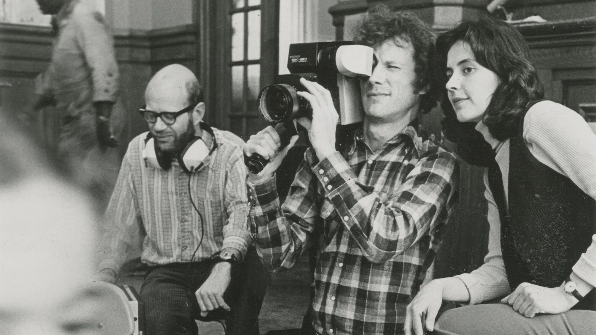 People sitting and watching a woman with a large camera in a room with wood paneling.