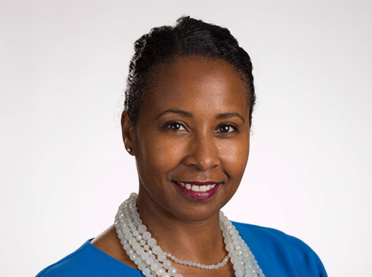 Portrait of a woman with short black hair wearing a blue top and a white pearl necklace, smiling against a plain white background.