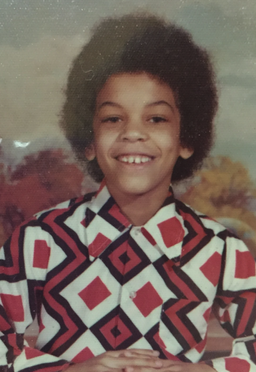 A young person with curly black hair, smiling and wearing a geometric patterned shirt with red, black, and white colors, sitting outdoors with trees in the background.