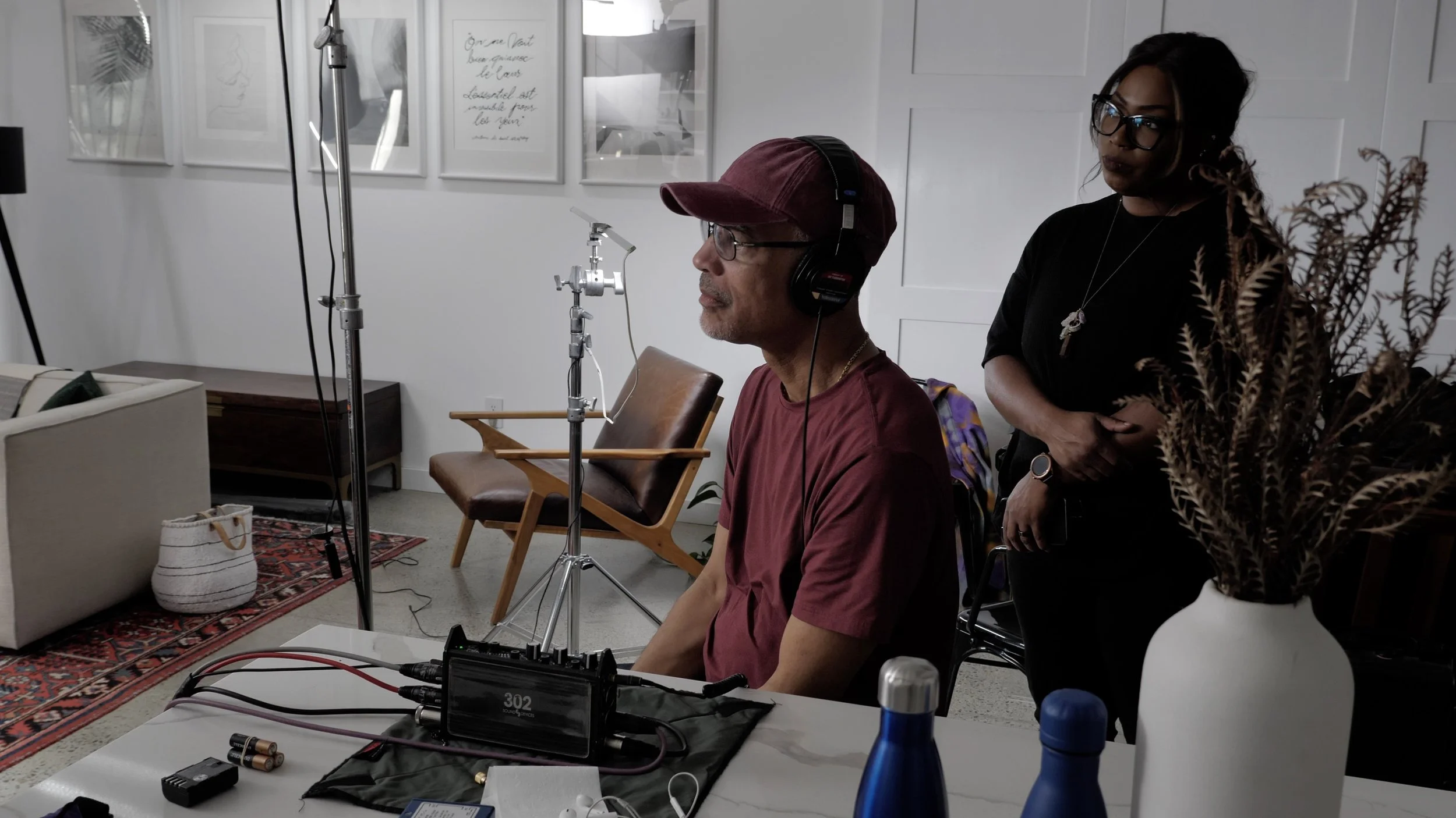 A man with glasses, a cap, and headphones sitting in front of audio equipment while a woman with glasses and a necklace watches. The setting appears to be a studio or living room with framed artwork on the wall, a leather chair, a patterned rug, and 