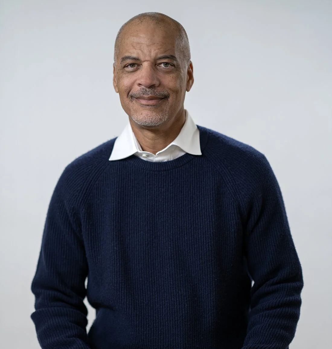 An older man with a bald head and gray beard, wearing a white collared shirt and a dark blue sweater, smiling softly at the camera against a plain light background.