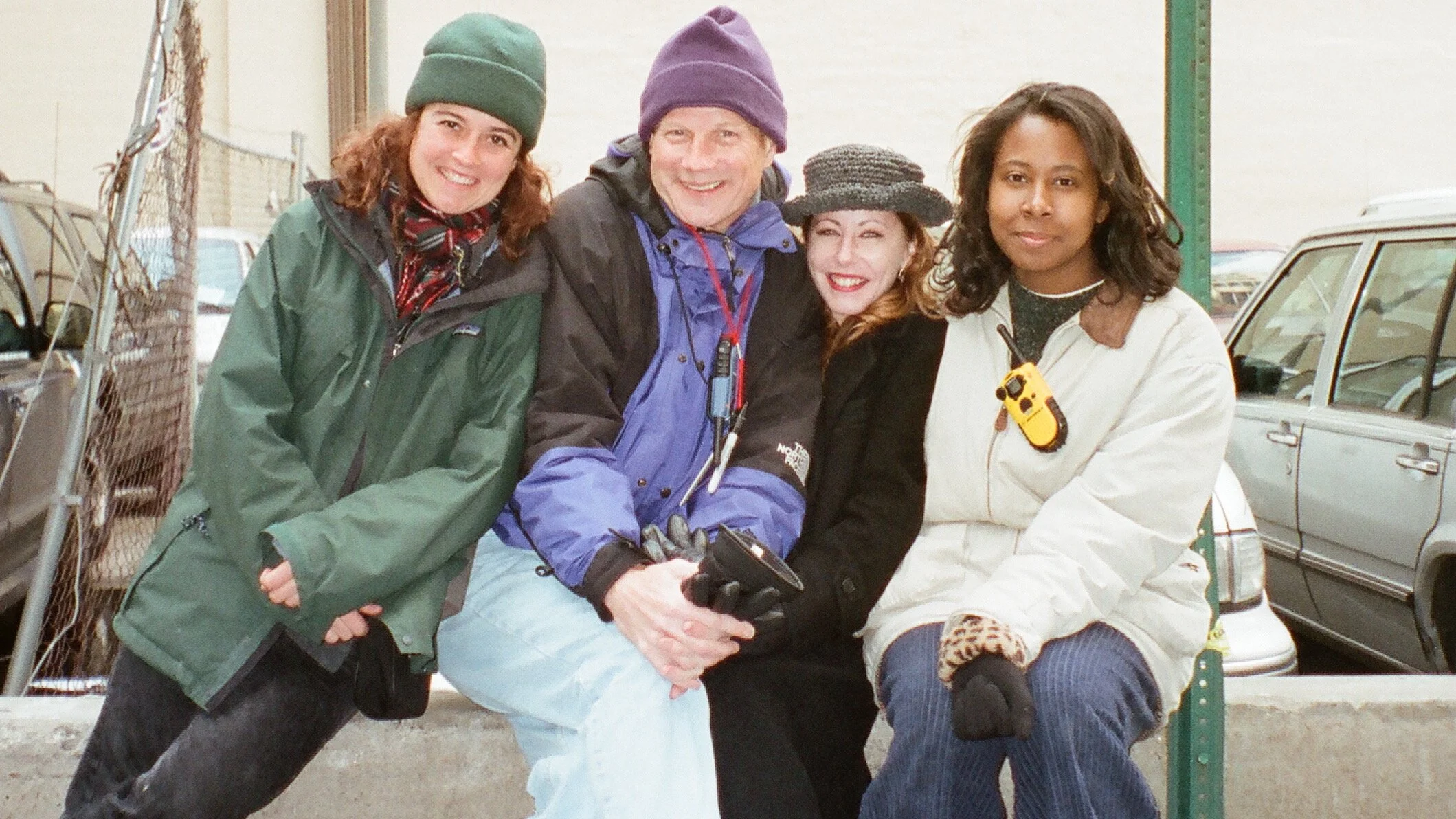 Four people posing together in an outdoor parking lot, dressed warmly in jackets and hats, with cars visible in the background.