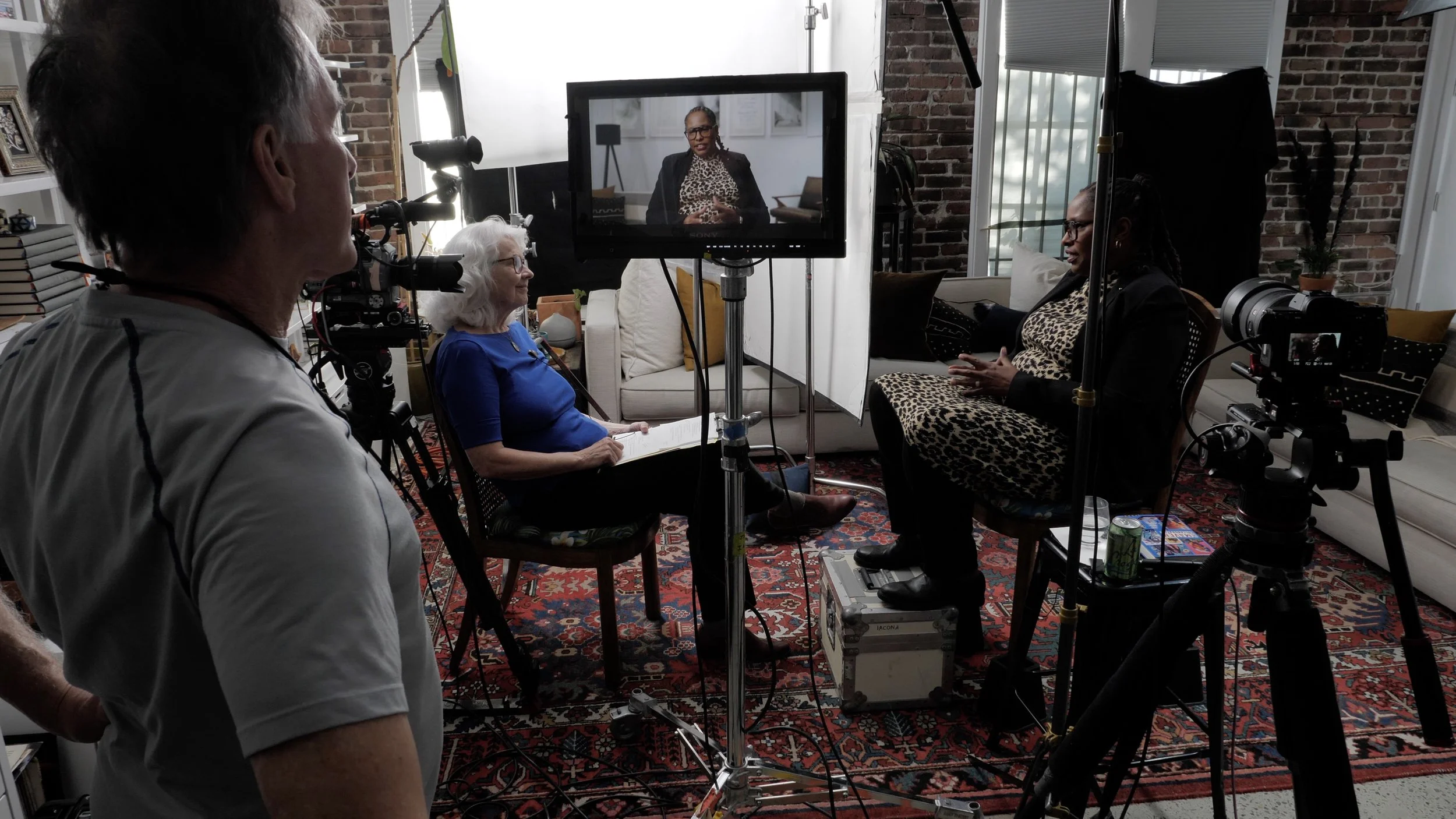 A woman in a patterned dress is being filmed during an interview in a living room with brick walls and large windows, with camera crew and equipment present.
