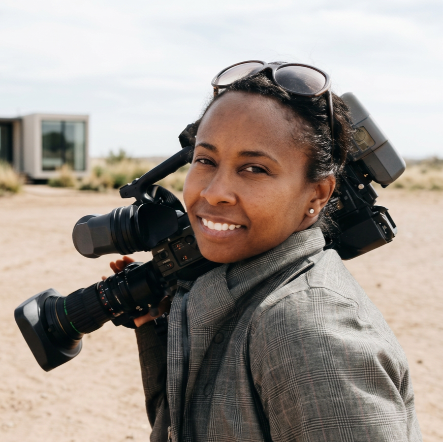 A woman smiling outdoors with a large camera on her shoulder, wearing sunglasses and a plaid jacket.