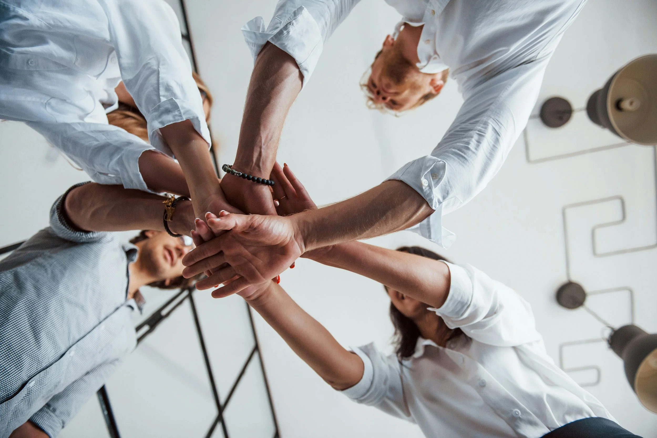 People in white shirts stacking hands together in a teamwork gesture in a meeting room.