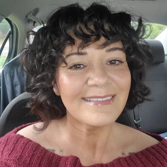 Close-up of a woman with curly dark hair, wearing a burgundy sweater, smiling inside a car.