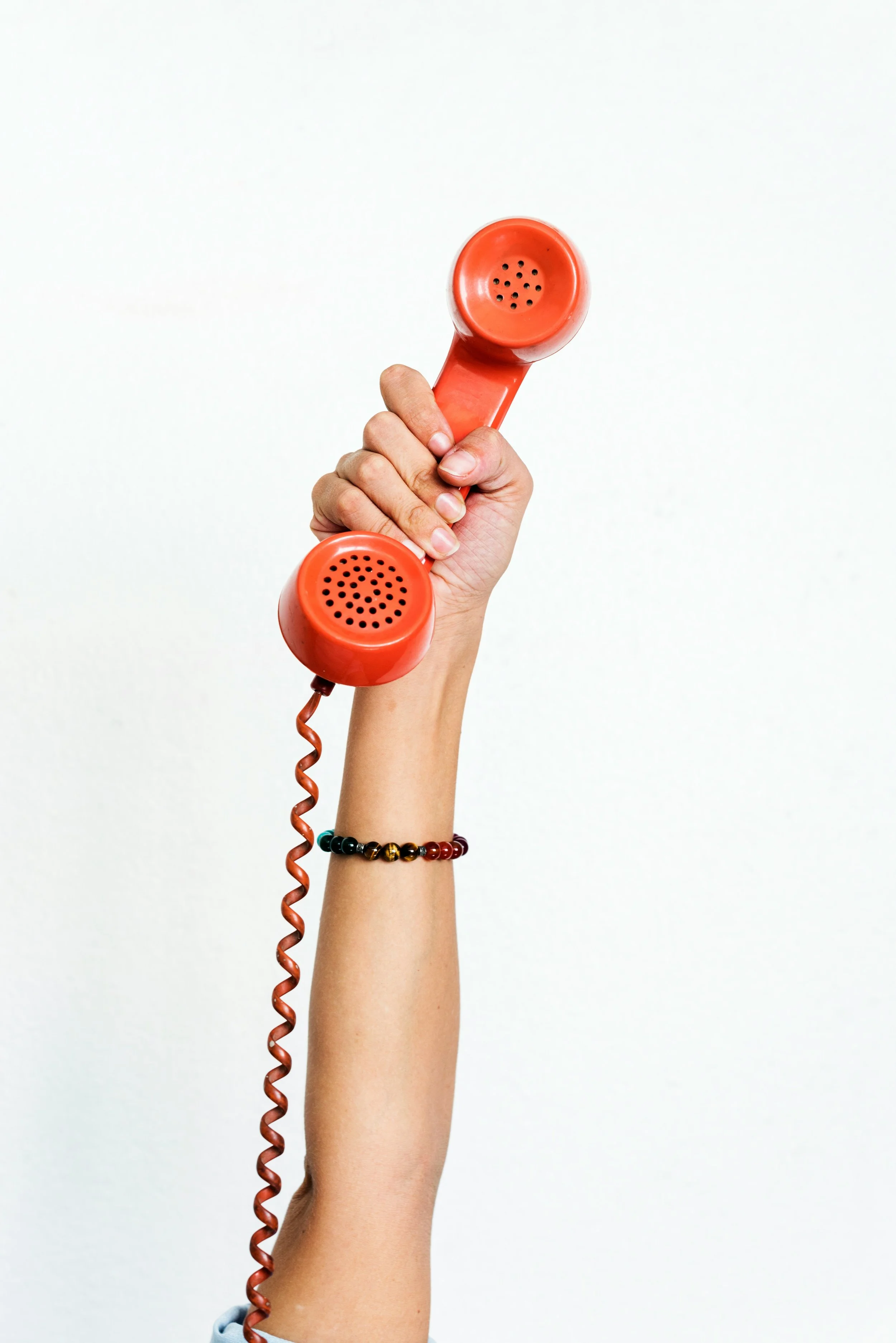 Person holding a red vintage telephone receiver against a plain white background, wearing a multicolored beaded bracelet.