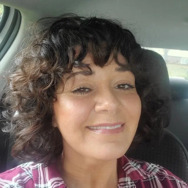 Close-up of a woman with curly dark hair and a septum piercing, smiling inside a car.