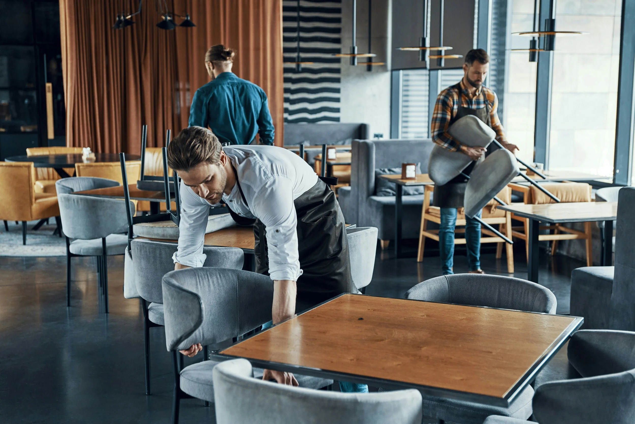 Three men cleaning and moving furniture in a modern restaurant with large windows, wooden accents, and contemporary furniture.