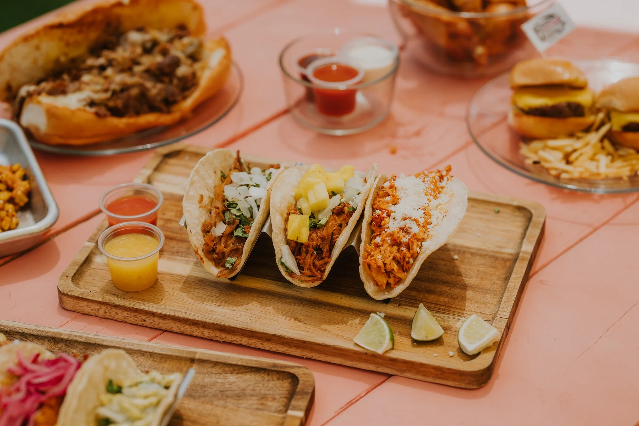 Three tacos filled with shredded meat, cheese, and vegetables on a wooden serving board, with lemon wedges and small cups of salsa and yellow sauce nearby, on a pink table.