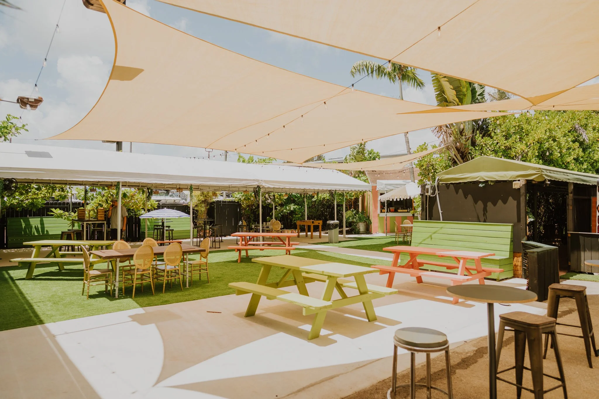 An outdoor patio area with colorful picnic tables, green cushioned benches, shaded by beige fabric canopies, surrounded by lush greenery and string lights.