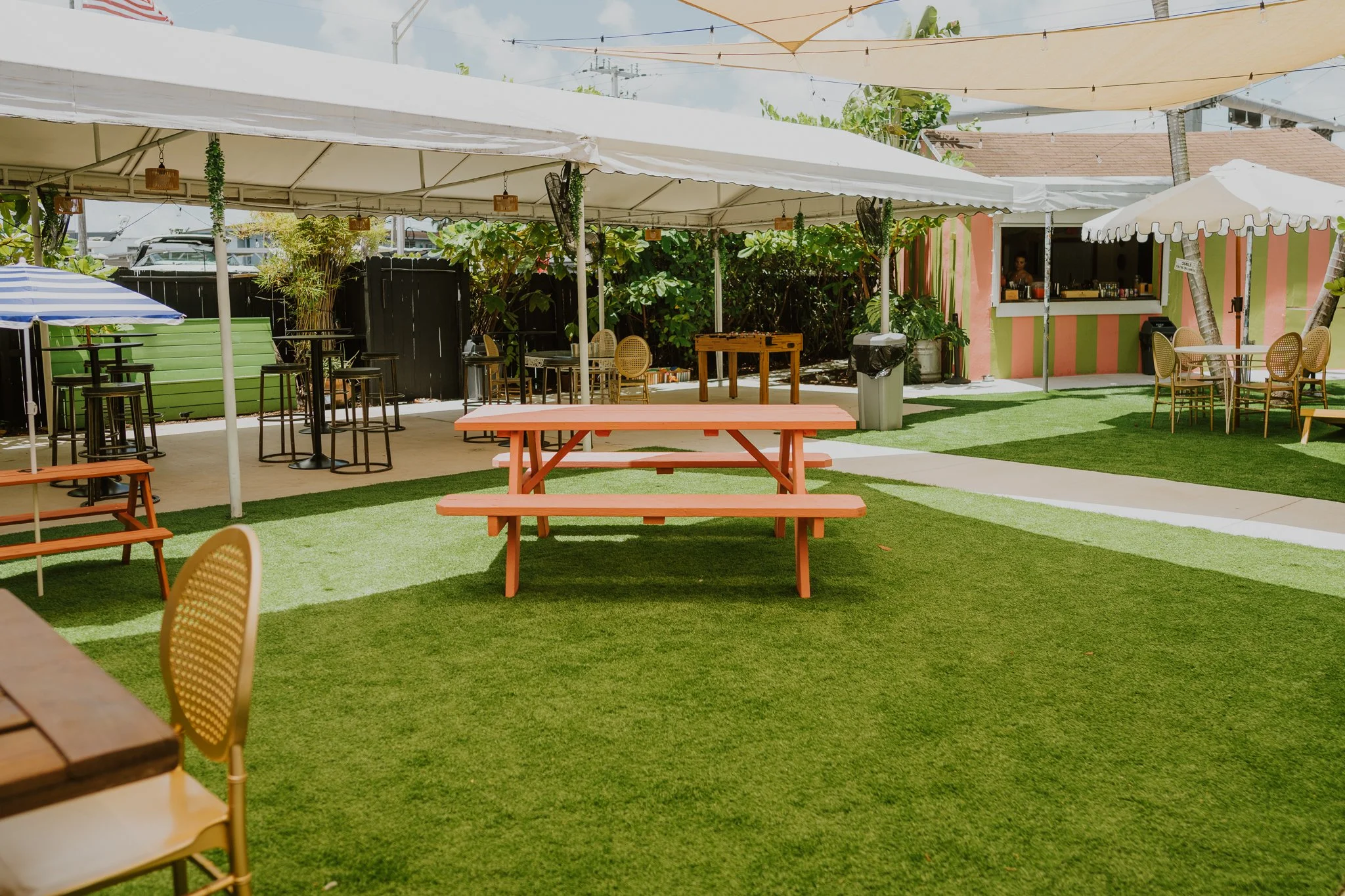 Outdoor patio area with pink and green striped wall, shaded seating area with tables and chairs, a serving window, umbrellas, and artificial grass.