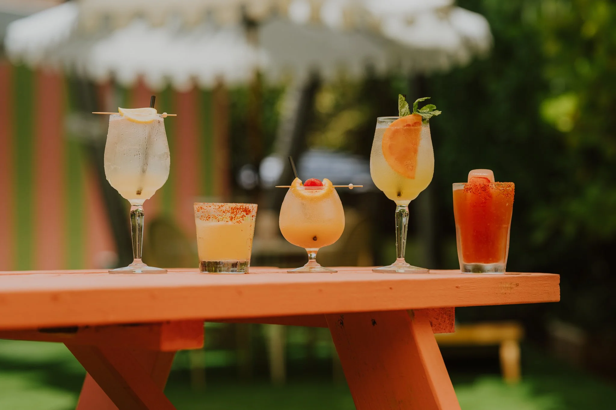 Various colorful cocktails and drinks with garnishes on a wooden table outdoors, with umbrellas and greenery in the background.
