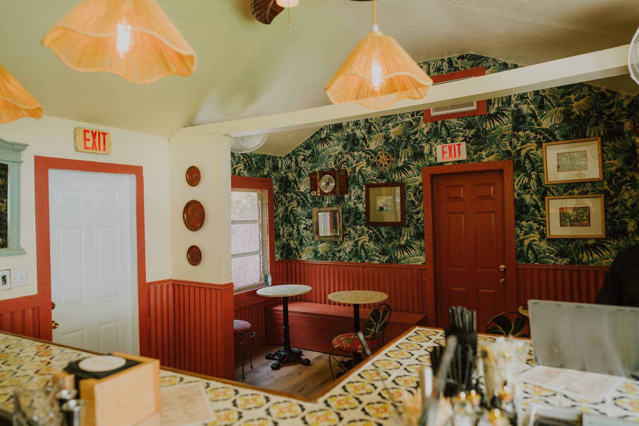 Interior of a cozy restaurant with tropical-themed wallpaper, wooden doors, framed artwork, hanging pendant lights, and small round tables with chairs.