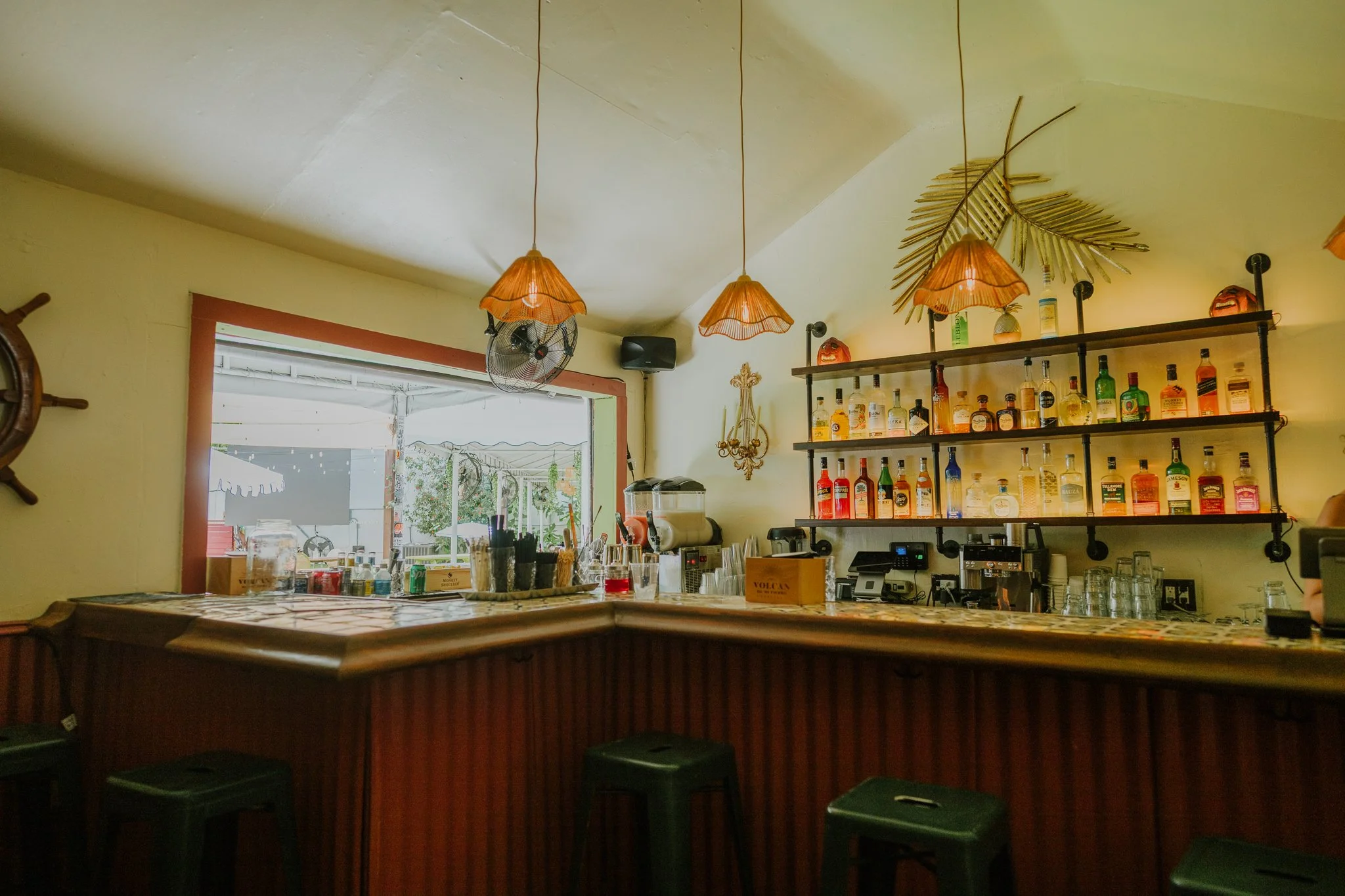A bar counter inside a restaurant with hanging lamps, a shelf with various bottles of alcohol, and an open window showing outdoor trees and a patio area.
