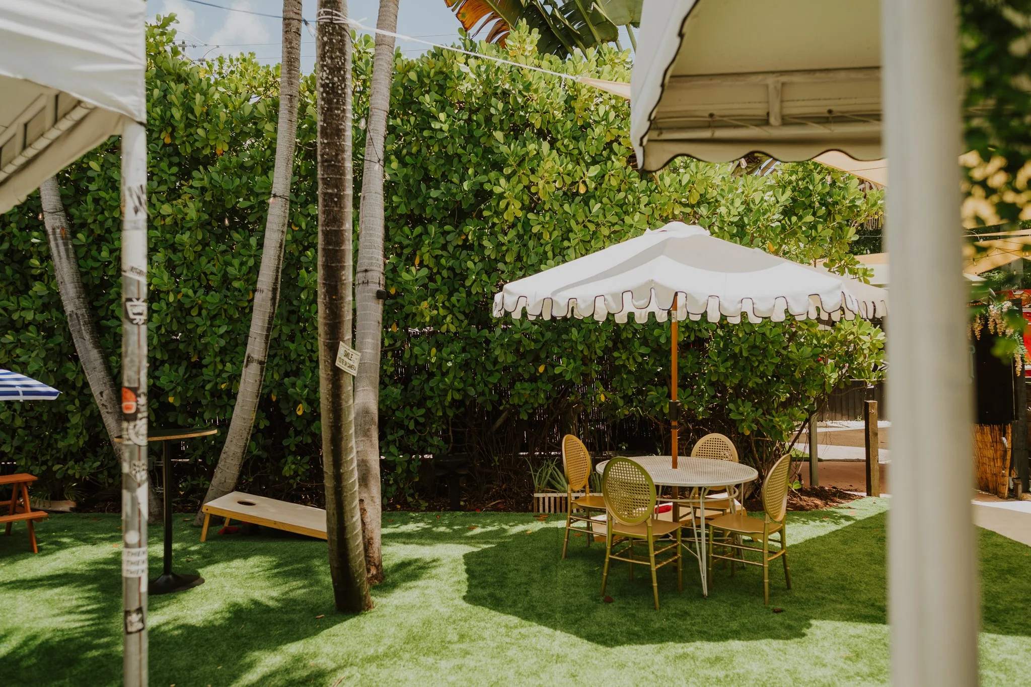 An outdoor patio with a round table and four chairs surrounded by lush green bushes and tall trees. Two white patio umbrellas are also visible, providing shade in the area.