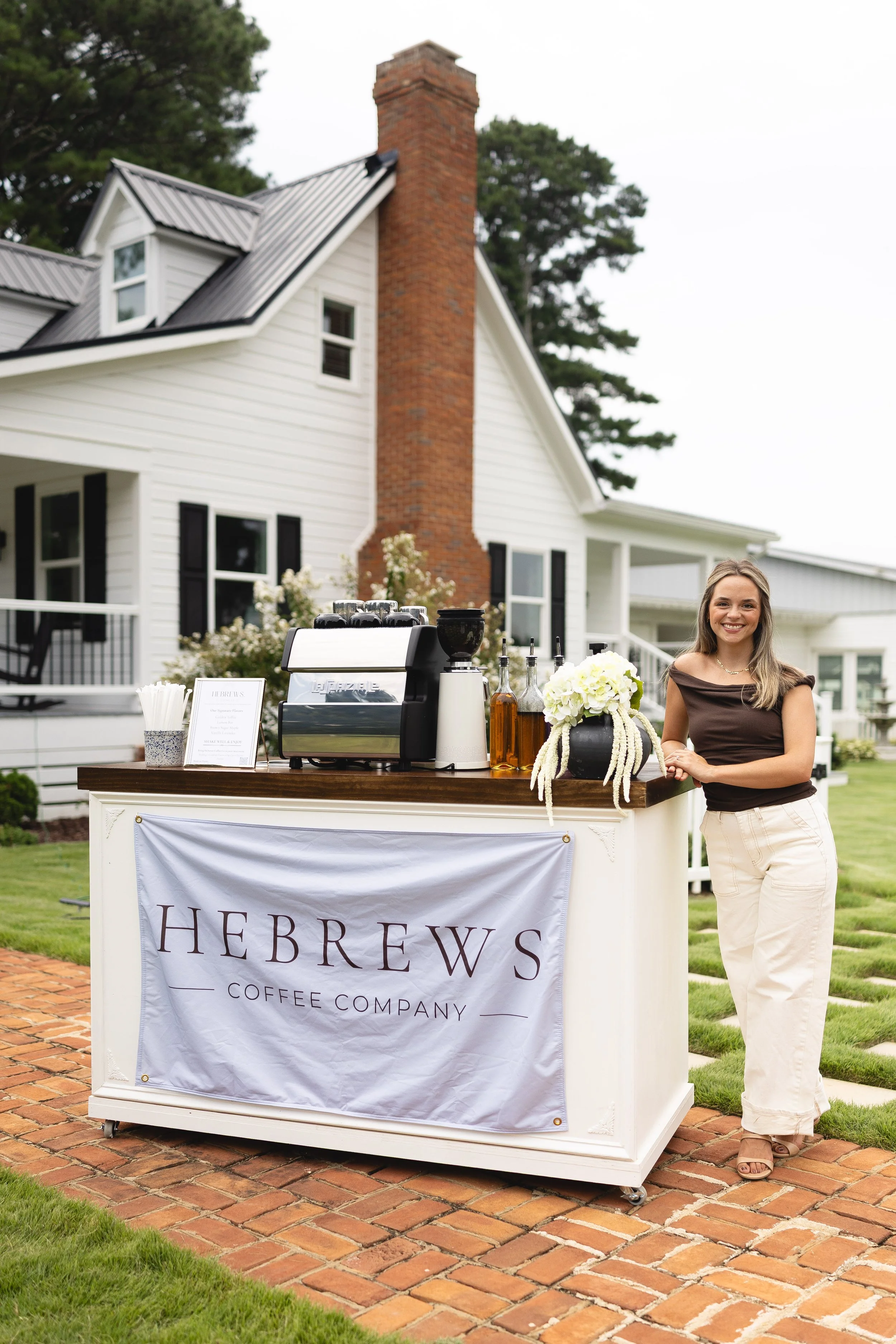 Hebrews Coffee Co. espresso cart with white florals set up outside a historic home for a wedding, with a barista ready to serve