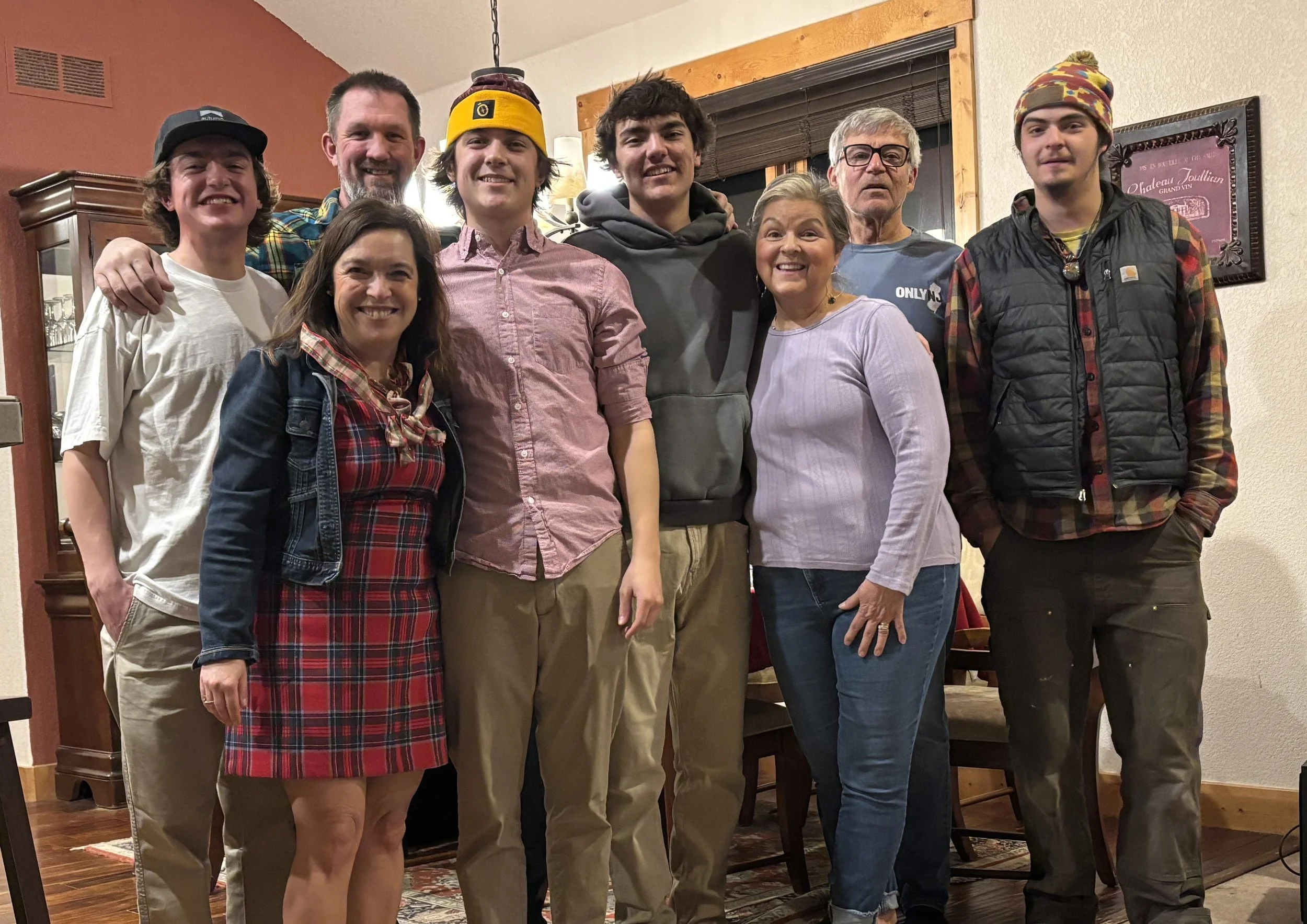 Nicole with her husband, sons, brother, and parents.  They are standing close together, posed for the picture, with a wooden wall, window, and framed art in the background.