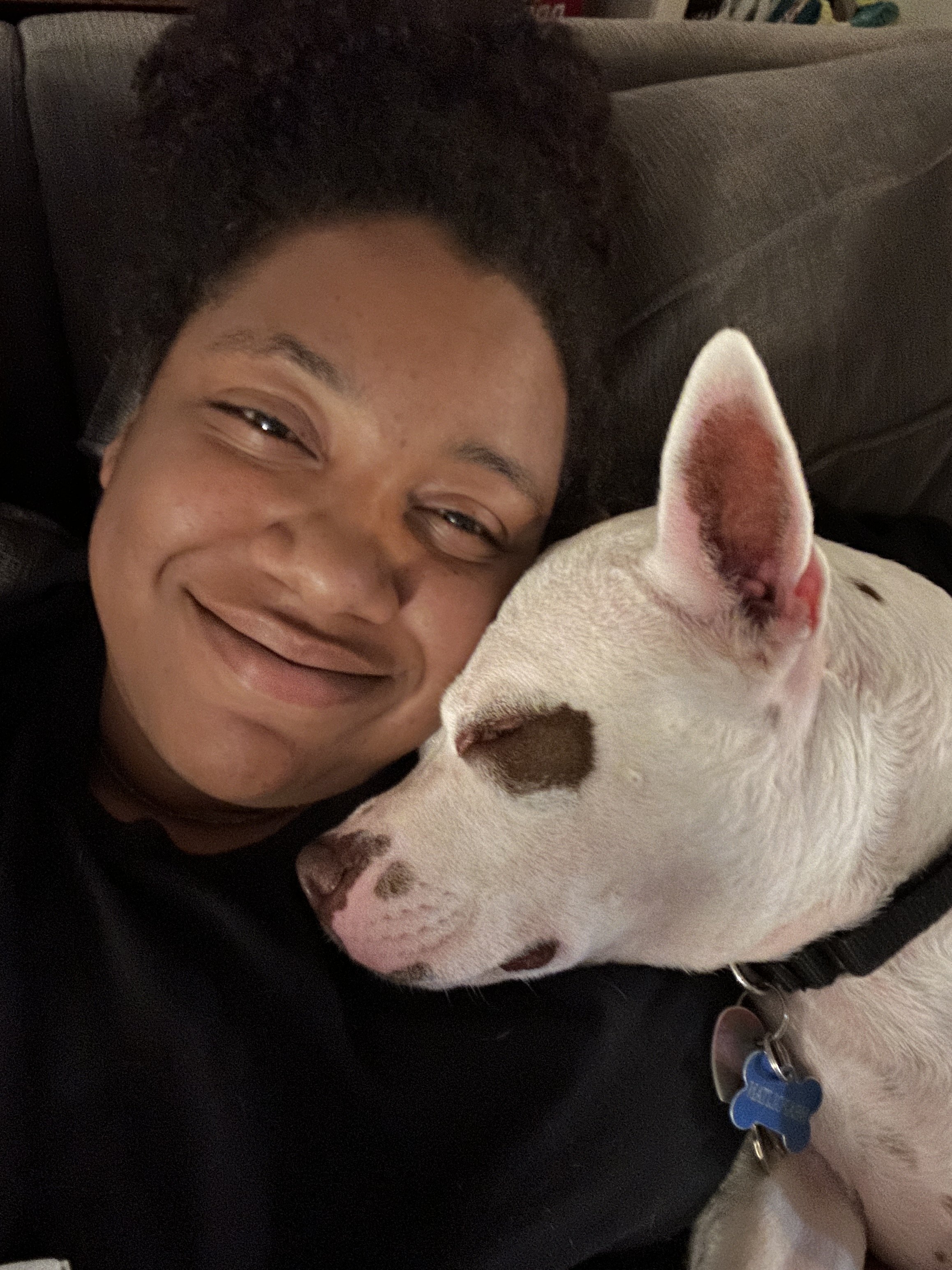 A woman and a white dog lying on a couch, close together. The woman is smiling and the dog has its head resting on the woman's arm.