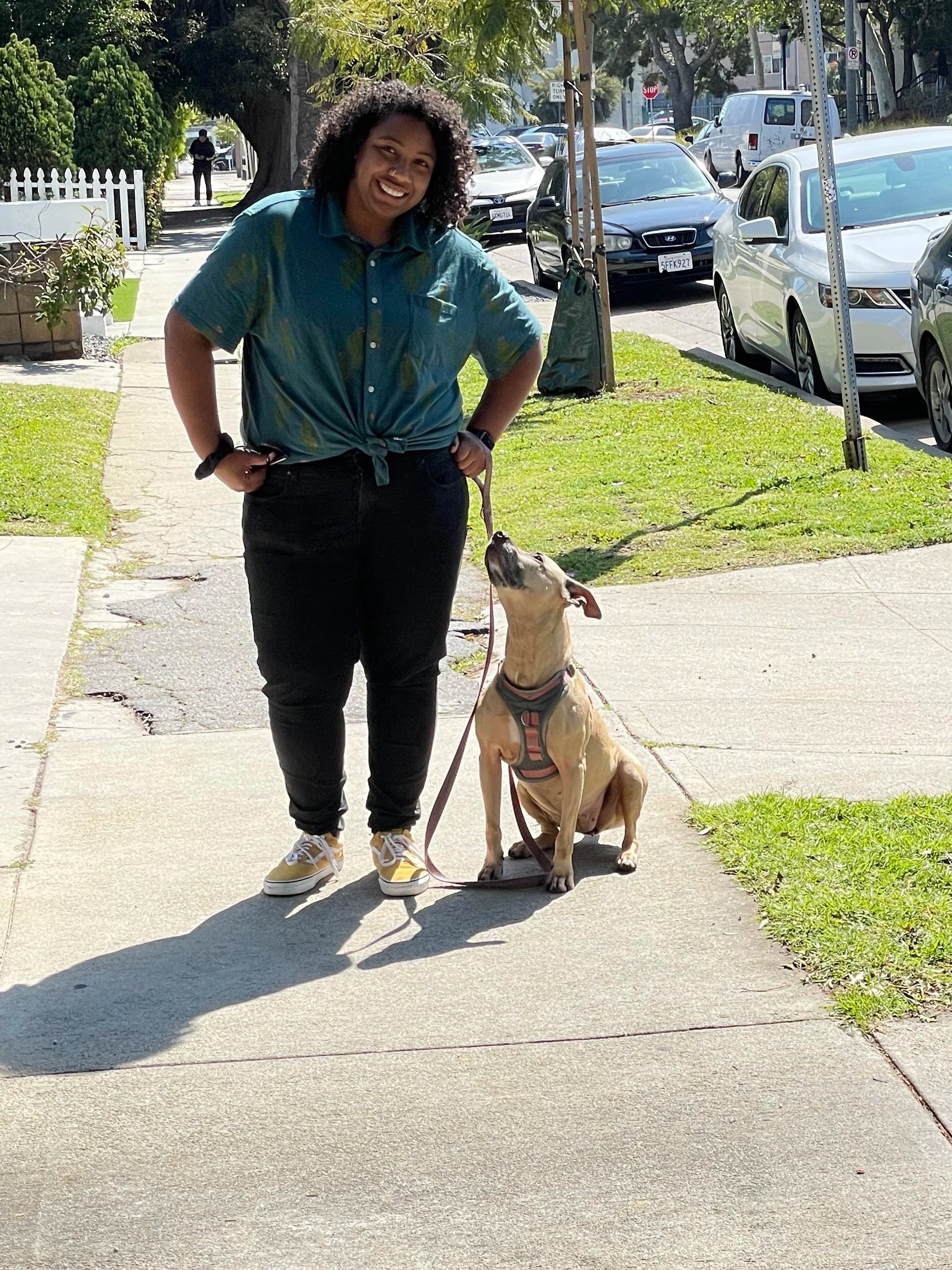 A woman with curly hair smiling and standing on a sidewalk, holding a leash attached to a large, tan-colored dog sitting on the grass. The woman is wearing a blue tie-dye shirt, black jeans, and yellow sneakers. There are parked cars, trees, and a person in the background.