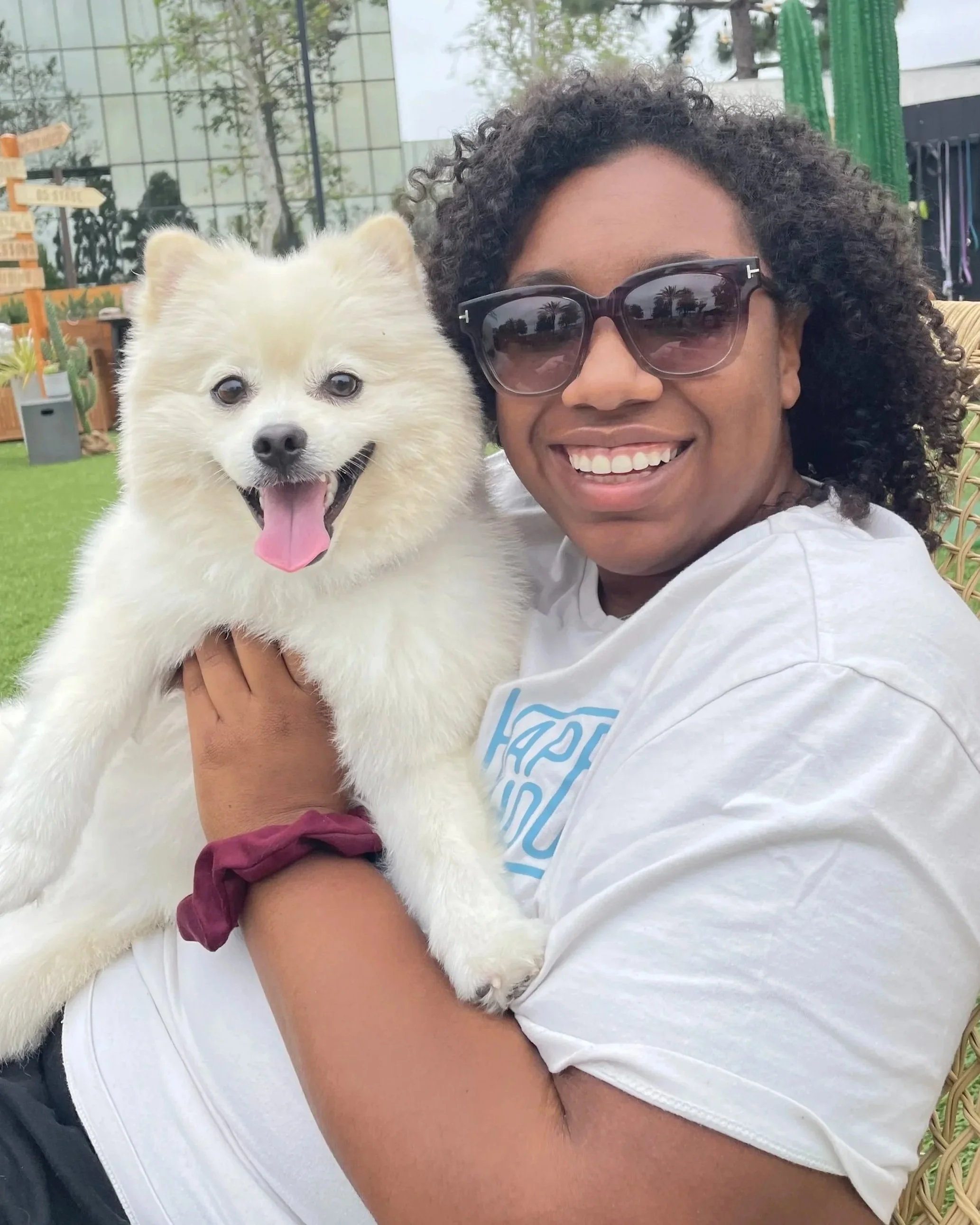 A woman with curly hair and sunglasses holding a fluffy white dog outside.