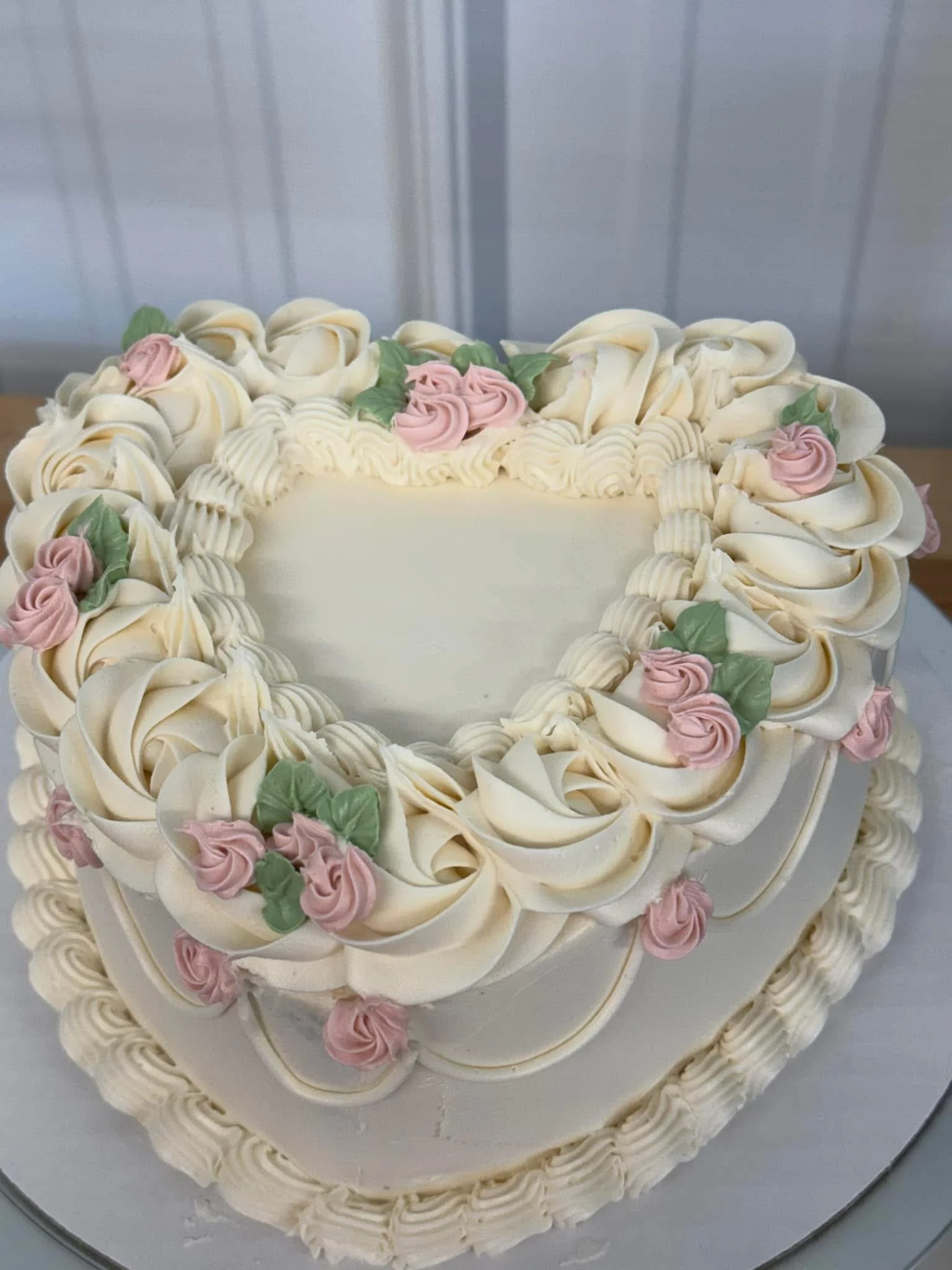 A heart-shaped cake decorated with white frosting, pink and green floral icing accents, and piped frosting border.