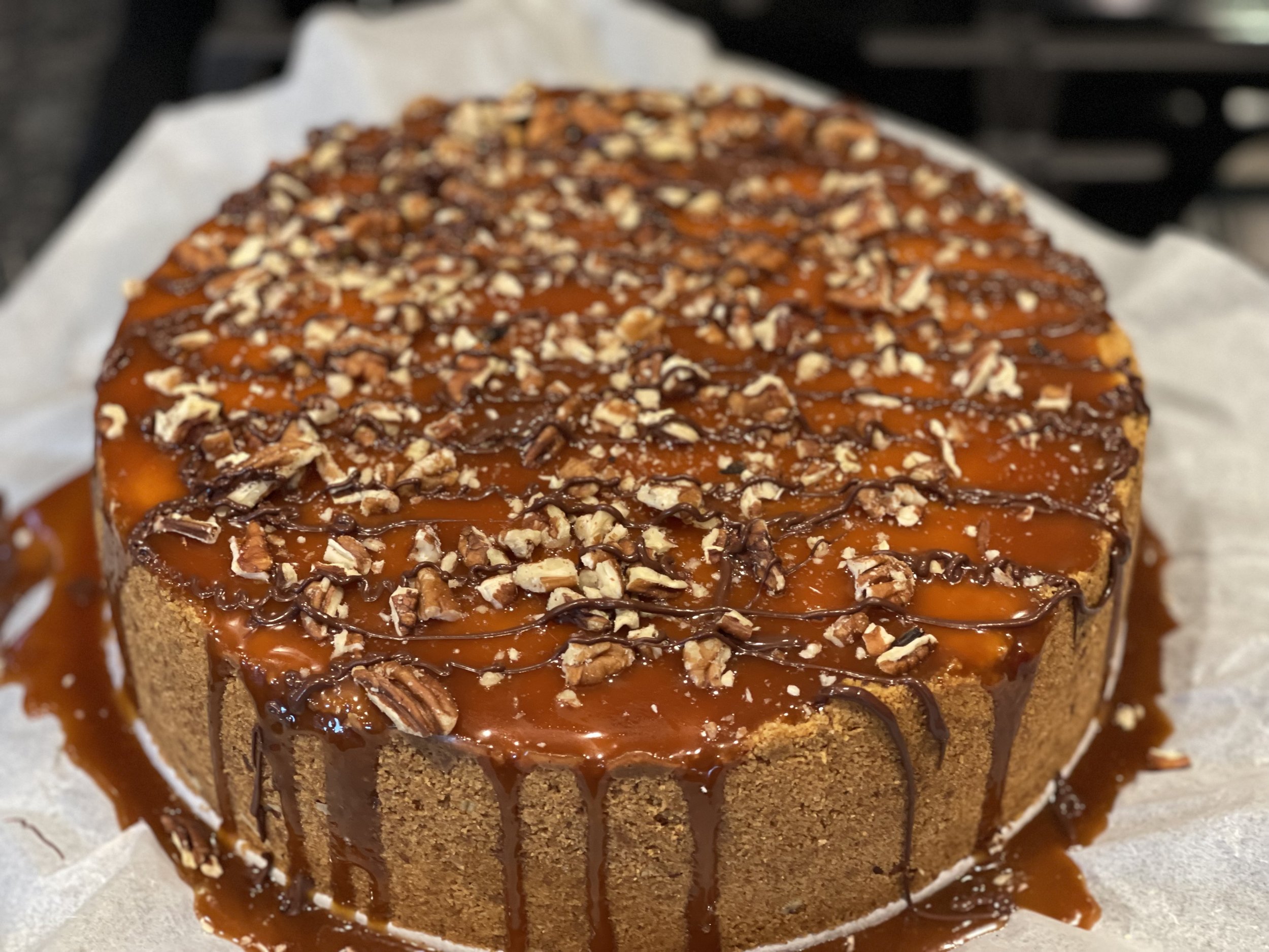 Close-up of a caramel cake topped with chopped nuts and drizzled with chocolate, sitting on parchment paper.