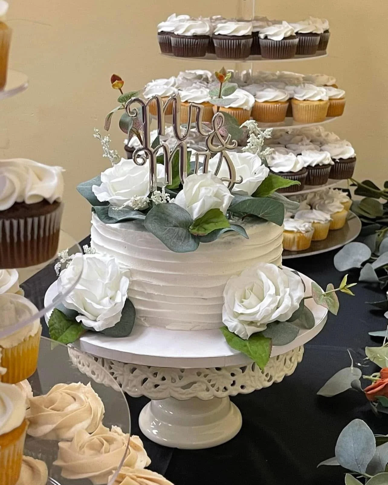 A wedding cake decorated with white roses and green foliage, topped with a "love" cake topper, surrounded by cupcakes with white frosting, on a decorative cake stand.