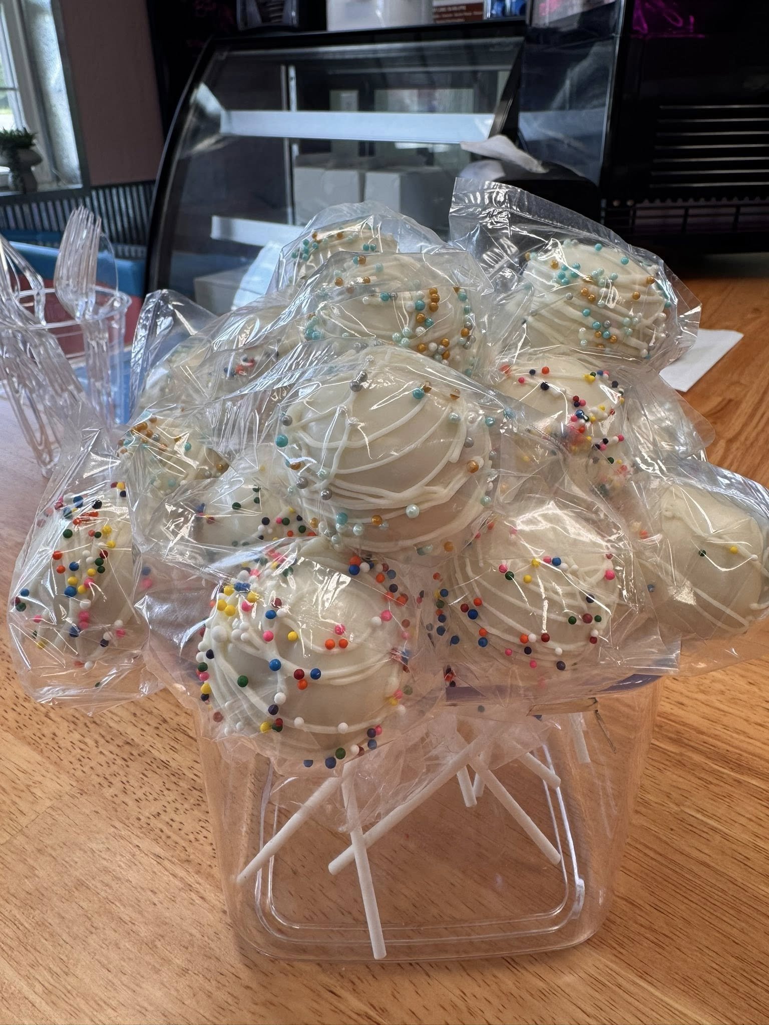 Several decorated cookies with white icing and colorful sprinkles, each individually wrapped in plastic, on top of white sticks. They are arranged on a wooden surface in front of a glass display case.