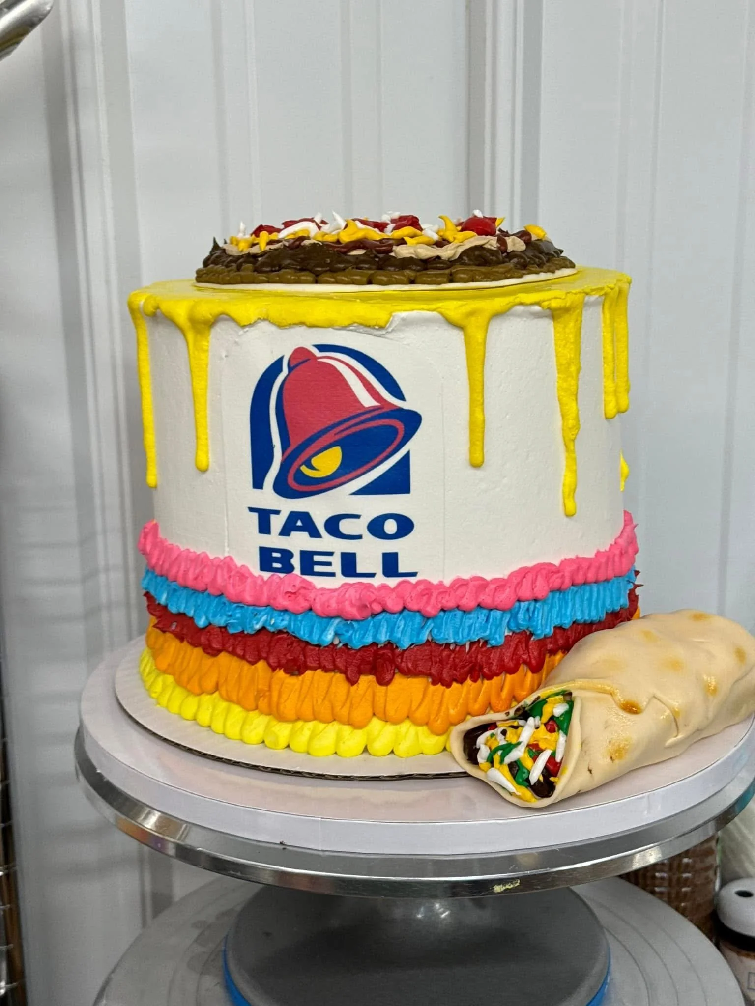 A colorful birthday cake with multiple tiers, decorated with yellow, pink, blue, and red icing, featuring the Taco Bell logo on the front, topped with chocolate and fruit, with a sprinkled churro next to it and a pastry in the foreground.