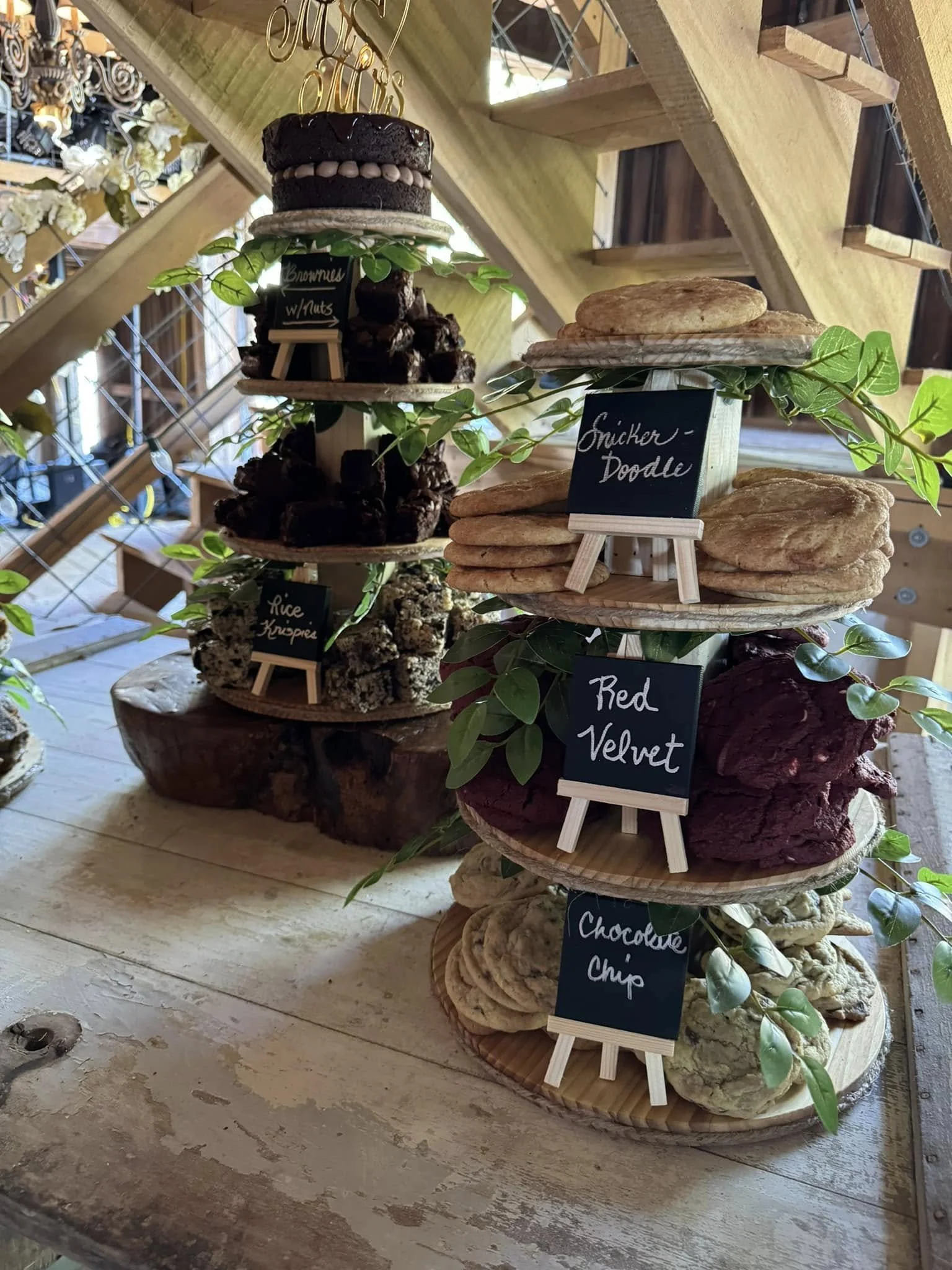 Display of various cookies on a wooden stand with small chalkboard signs indicating flavors like snicker-doodle, red velvet, and chocolate chip. Green leaves decorate the display.