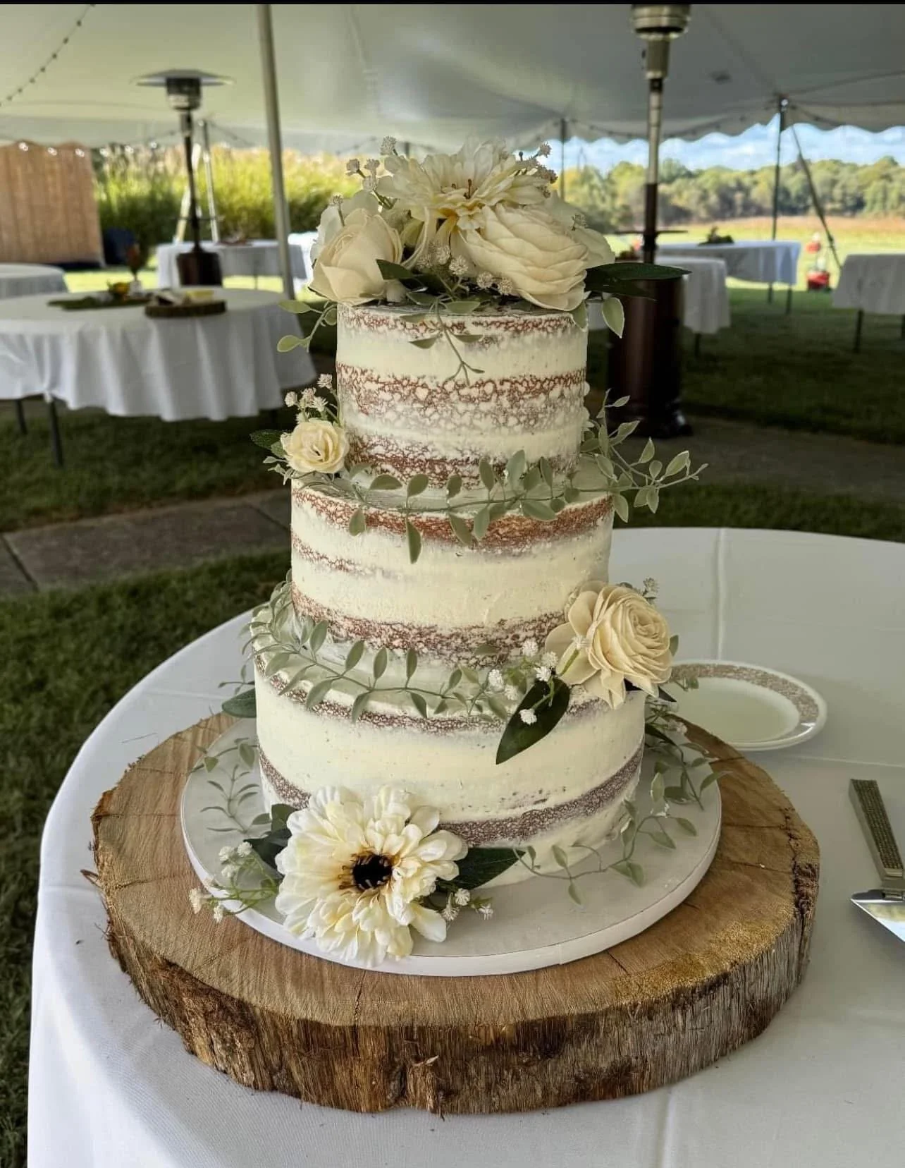 A semi-naked wedding cake with white frosting and brown cake layers, decorated with white roses, green leaves, and small white flowers, placed on a wooden slab on a table.
