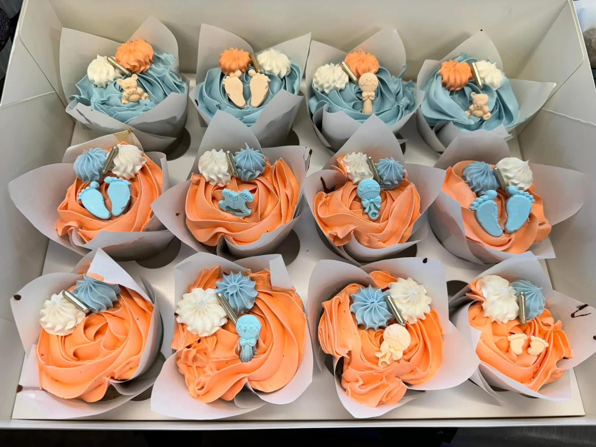 A box of cupcakes decorated with orange, blue, and white frosting, topped with small fondant baby-themed decorations and charms, each cupcake wrapped in white paper.