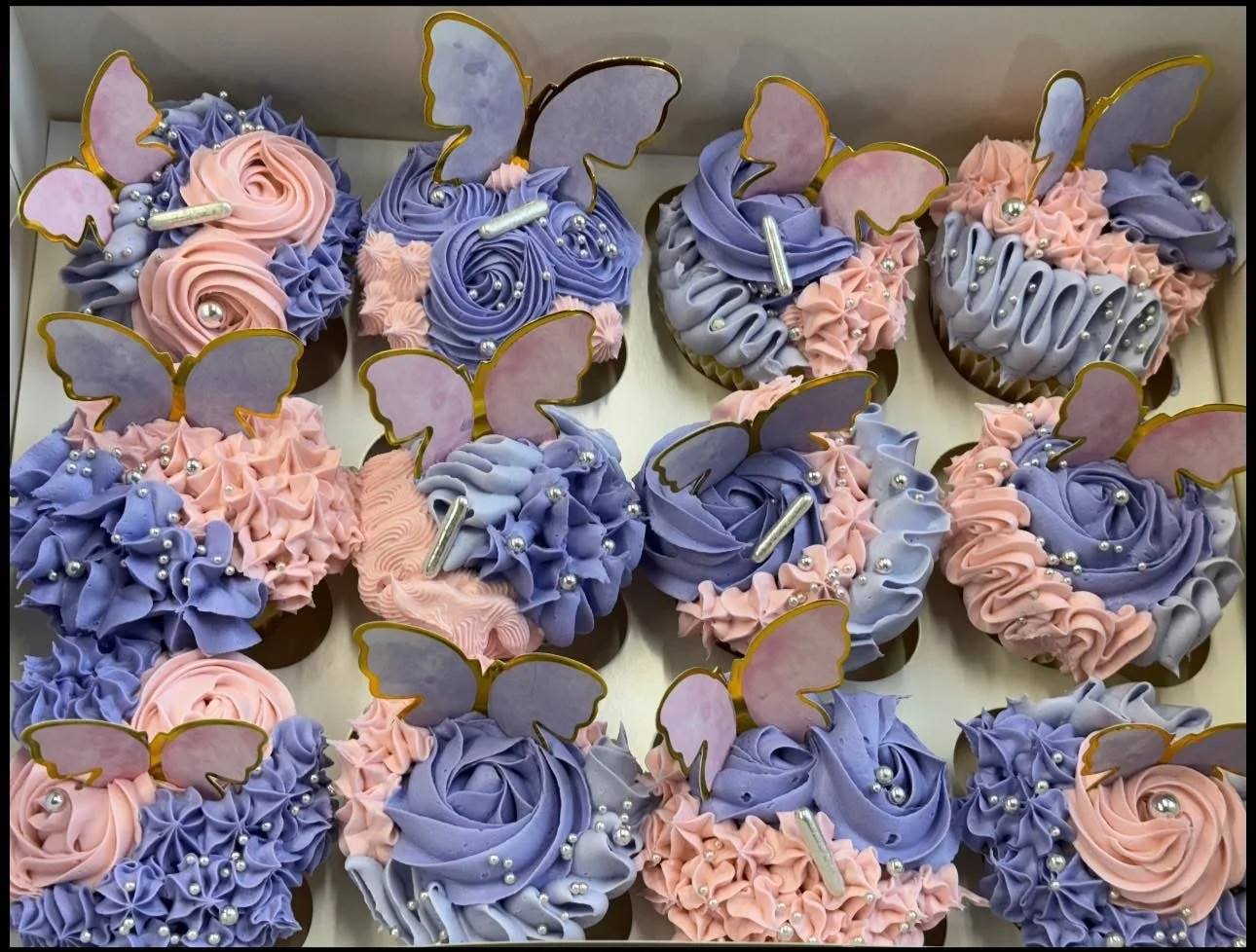 A box of decorative cupcakes with purple and pink frosting, each topped with edible butterfly and silver sugar pearls.