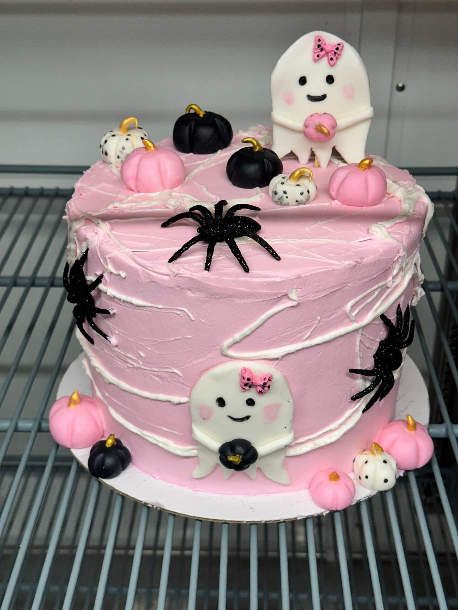 Pink Halloween cake decorated with white and black spider, ghost-shaped fondant with pink bow, pumpkin-shaped confections, and white ghosts with pink bows on top.