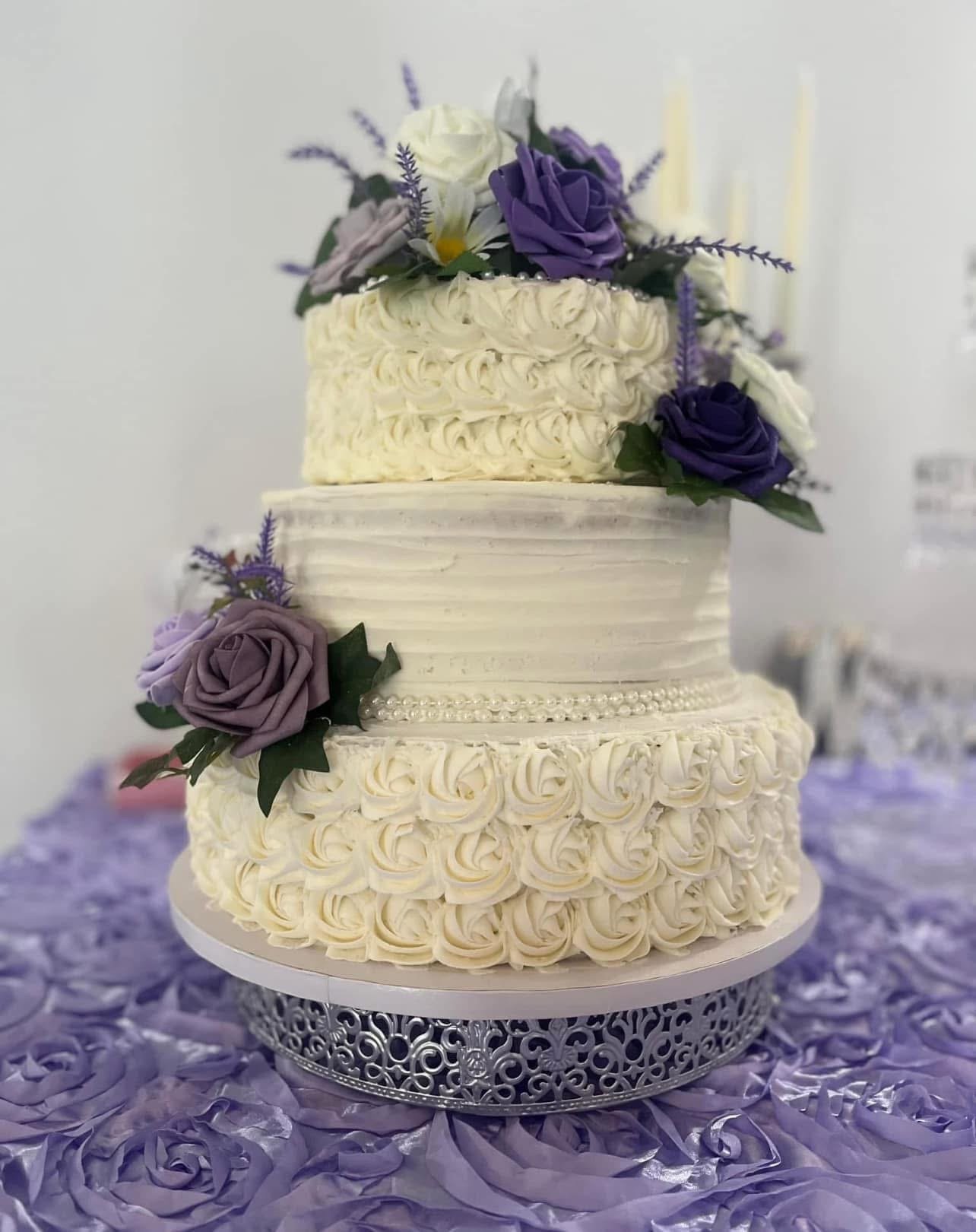 Three-tiered wedding cake decorated with purple and white roses, lavender, and pearl accents, with the top tier topped with flowers and pearls, on a silver cake stand on purple petal-like table covering.