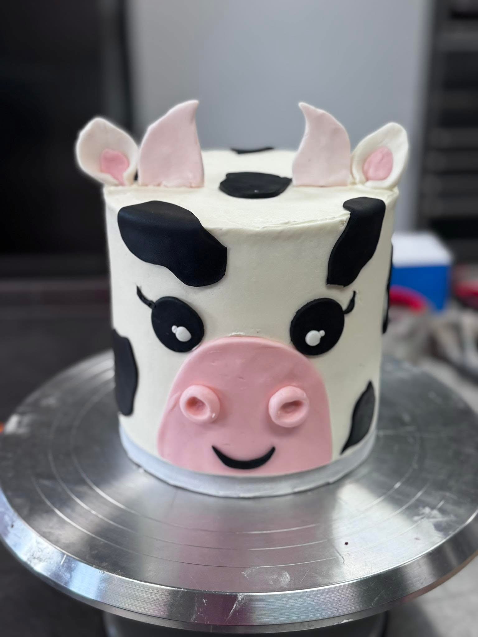 A cake decorated to look like a cow, with fondant ears, eyes, nose, and black spots, placed on a metallic surface.