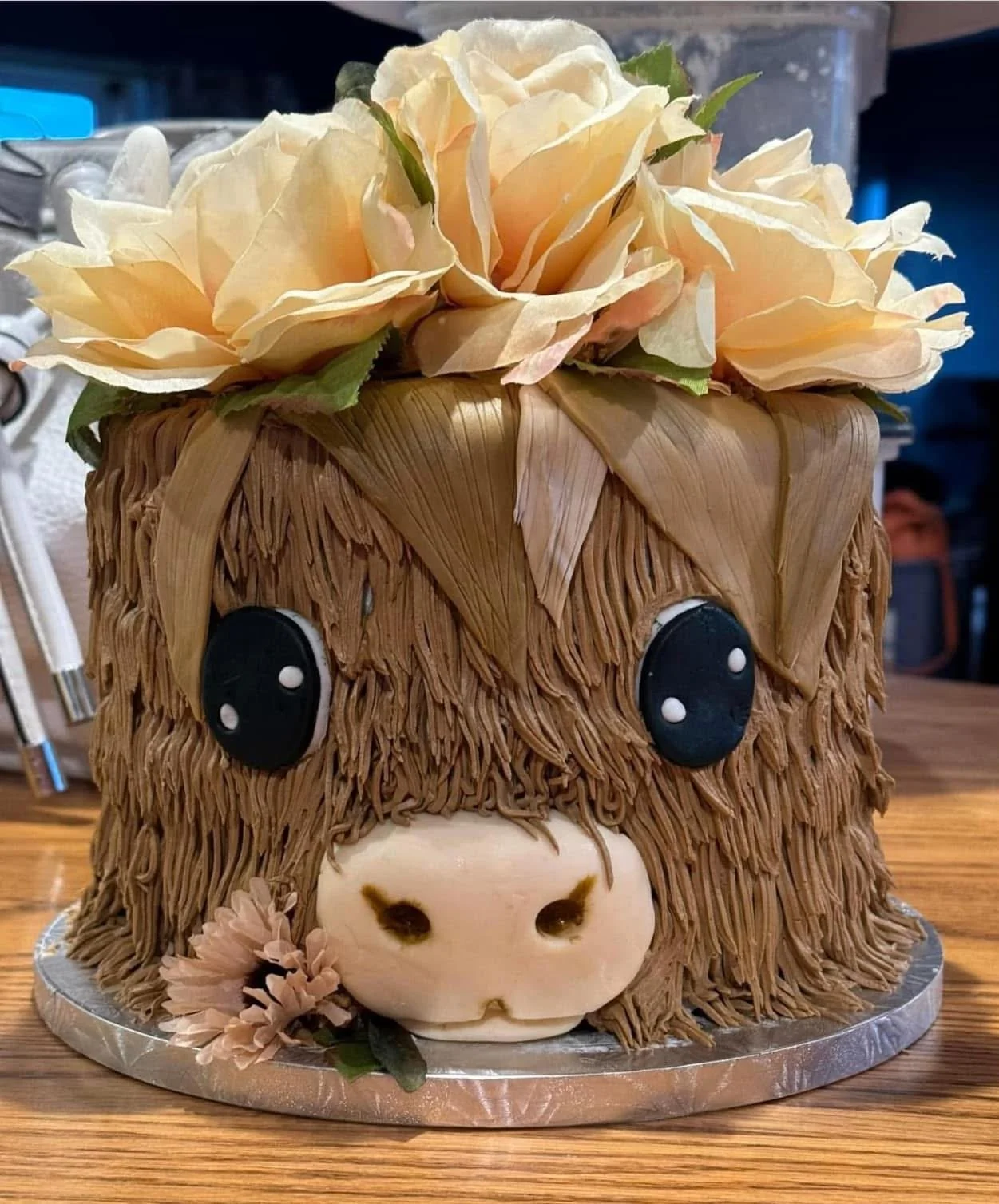 Cake decorated to look like a horse's face with beige and pink sugar flowers on top, brown textured frosting resembling horse hair, black fondant eyes with white dots, and a white fondant snout with nostrils, placed on a round silver cake board.