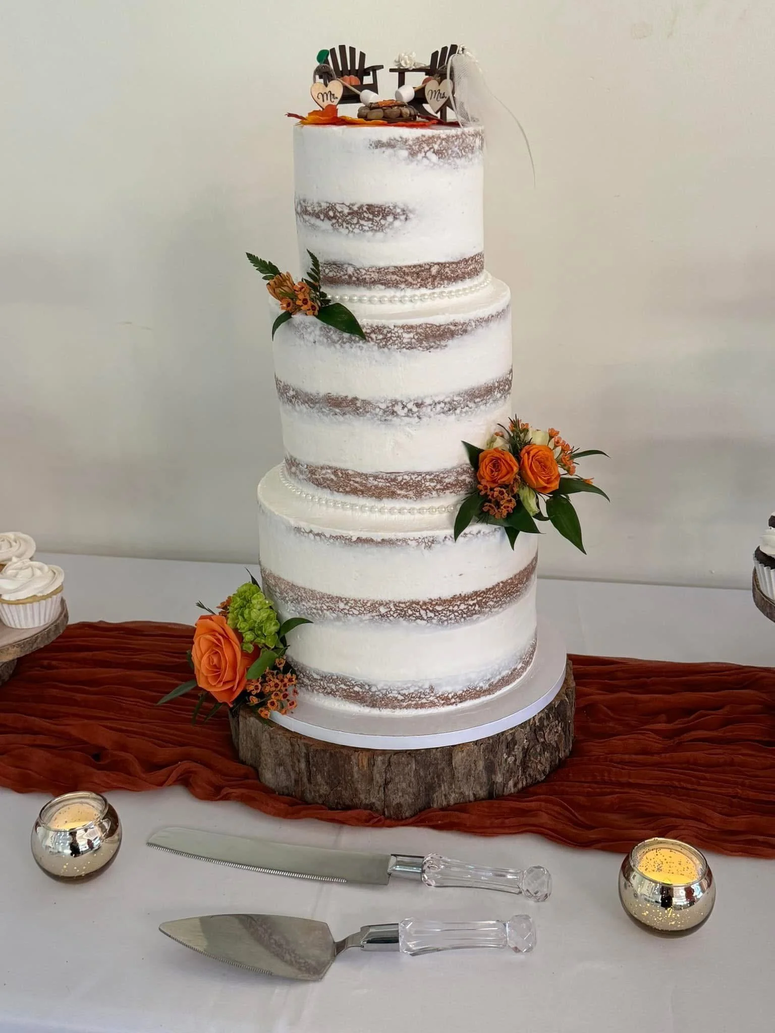 Three-tier rustic wedding cake with white and brown frosting, decorated with flowers and greenery, placed on a wooden slab. The background shows a table with a white tablecloth, surrounded by small candles and wedding cake serving utensils.
