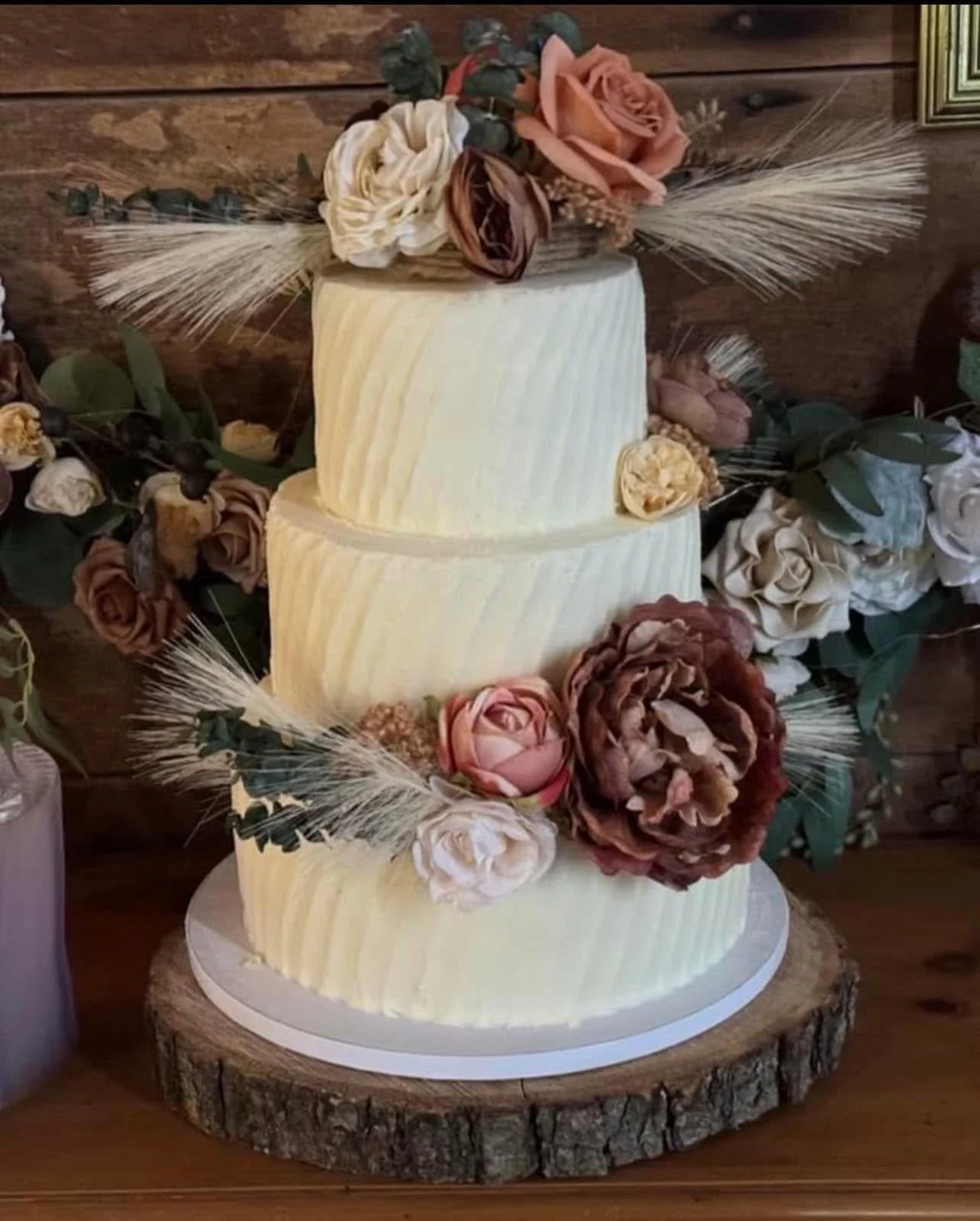 Three-tiered wedding cake decorated with dried roses, peonies, and wheat stalks, placed on a wooden slab, with floral arrangements surrounding it.