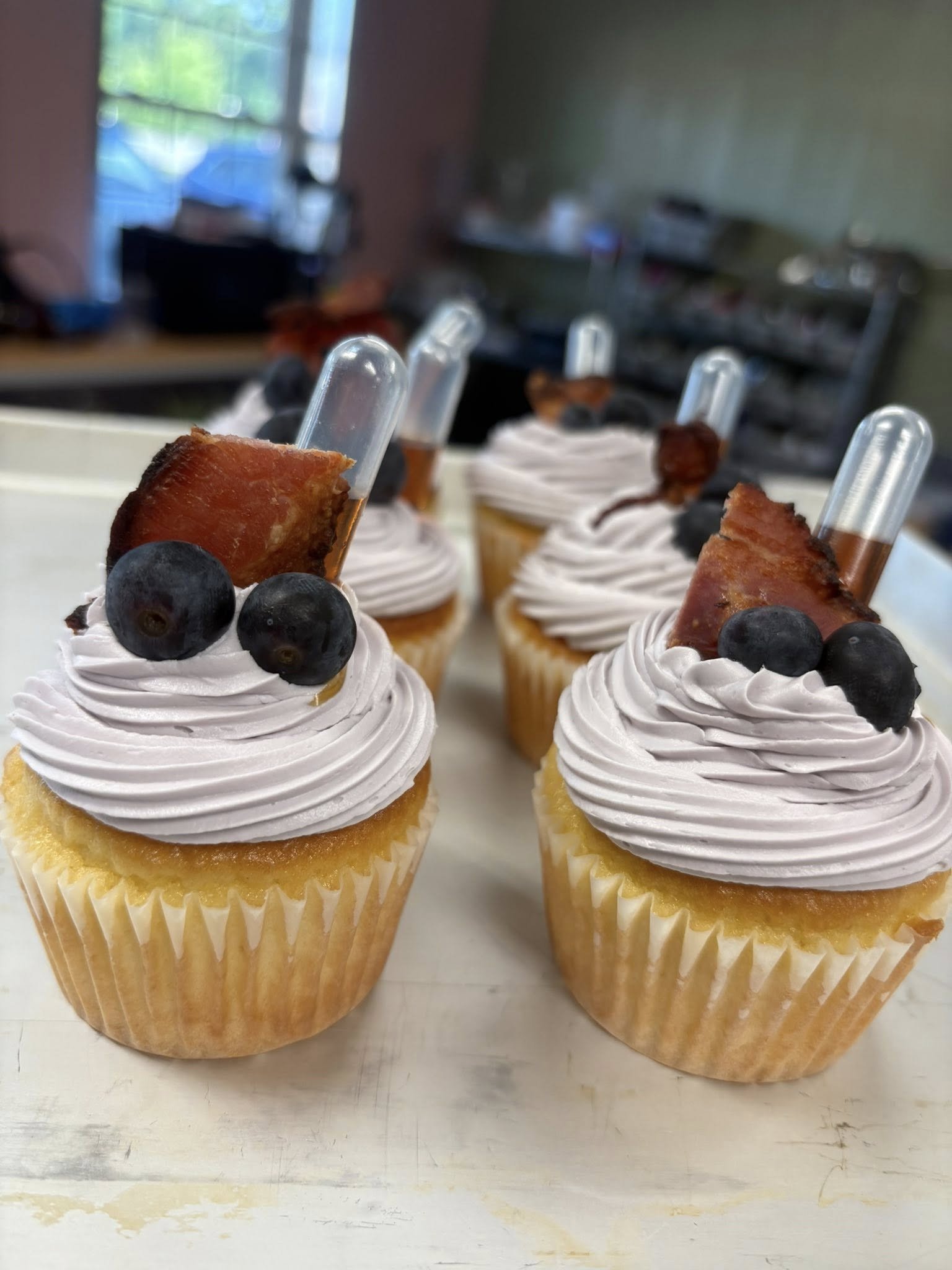 Close-up of four cupcakes decorated with pinkish-purple swirled frosting, topped with blueberries and pieces of bacon, each with a clear plastic tube inserted, on a white surface with a blurred kitchen background.