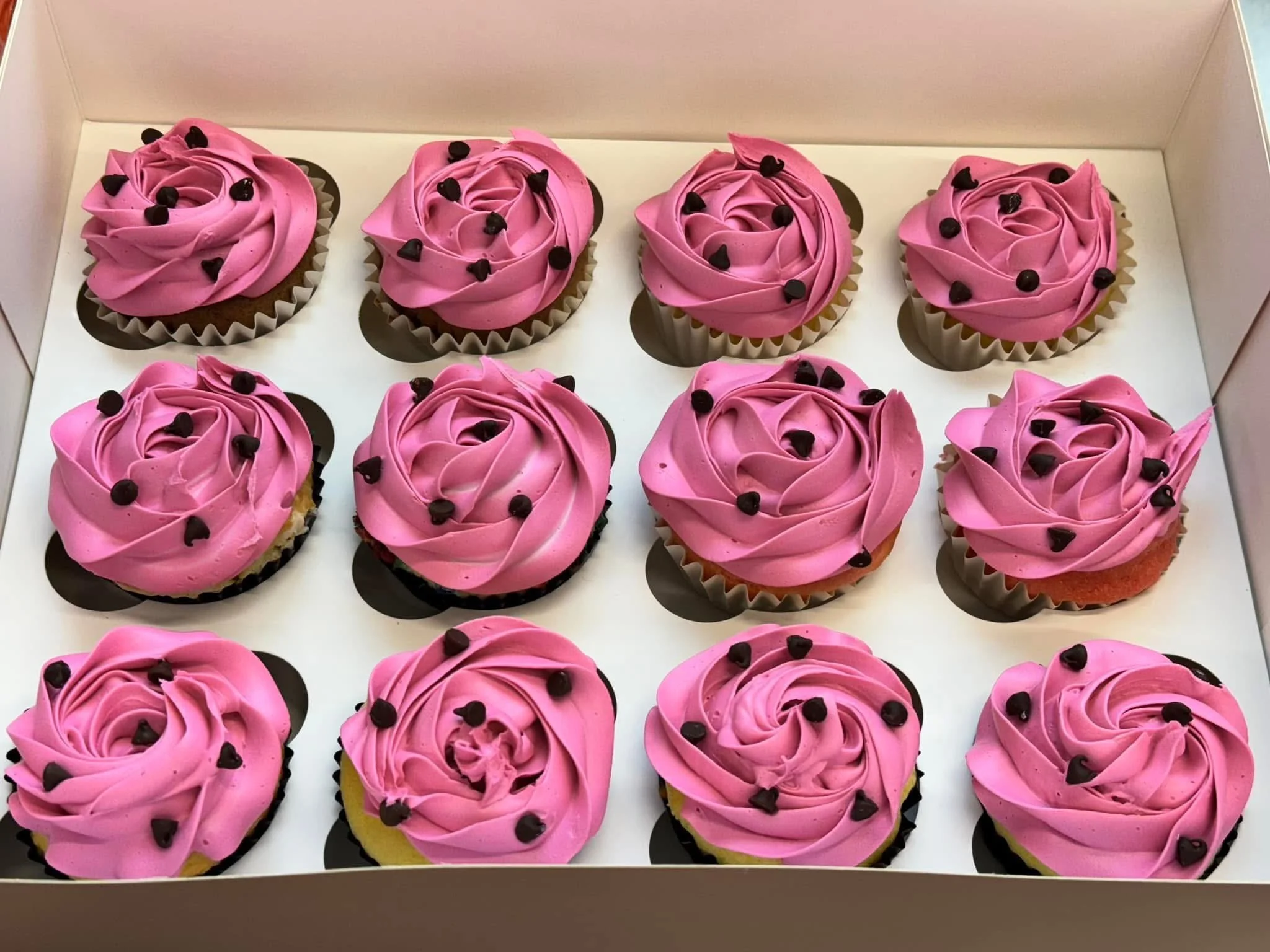 A white box contains twelve cupcakes with pink frosting and small black heart-shaped candies on top, arranged in three rows of four.