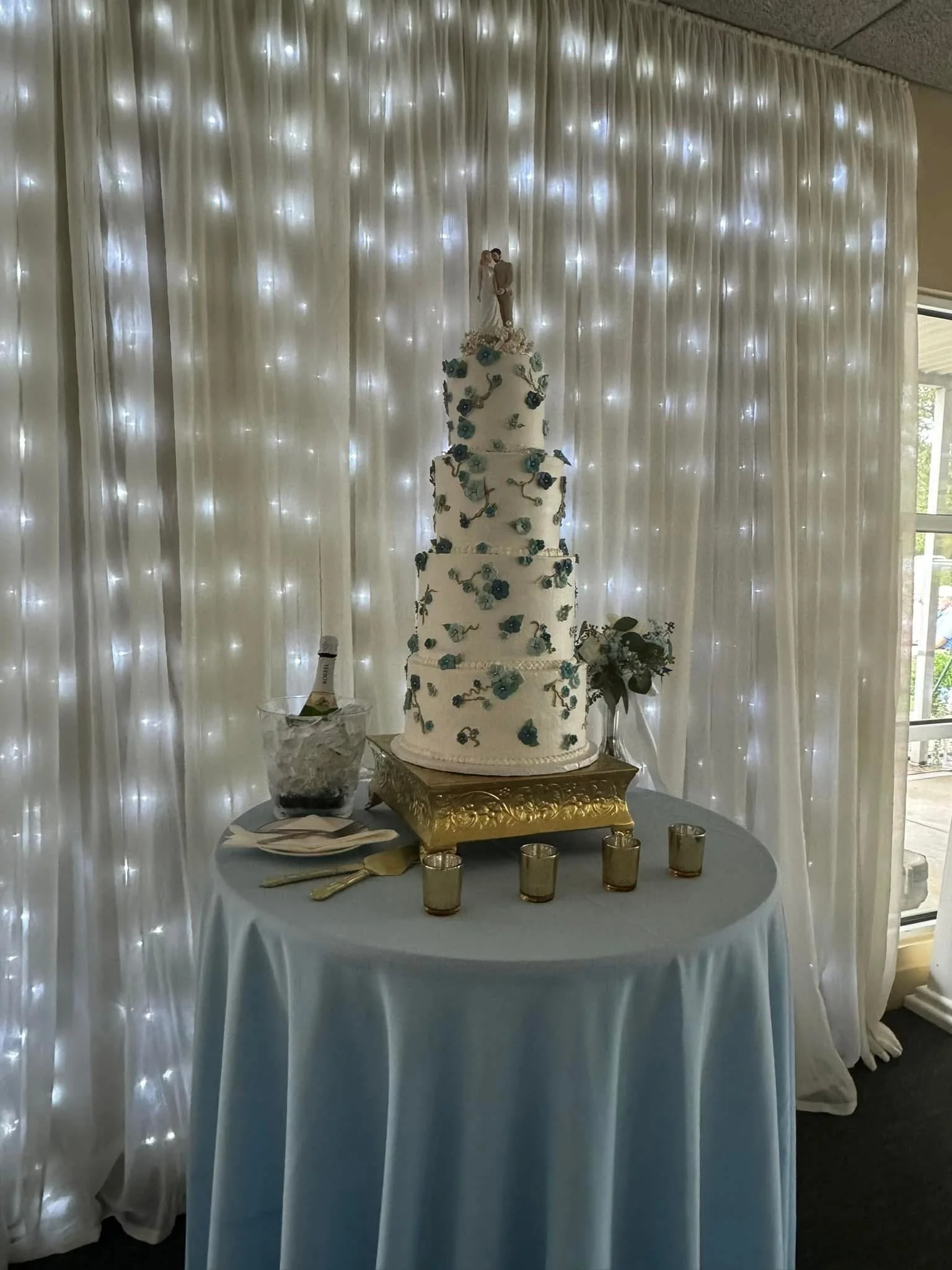 Elegant four-tier wedding cake decorated with blue flowers and green vines, topped with a bride and groom figurine, displayed on a table with gold candles, set against a backdrop of white curtains with twinkling lights.
