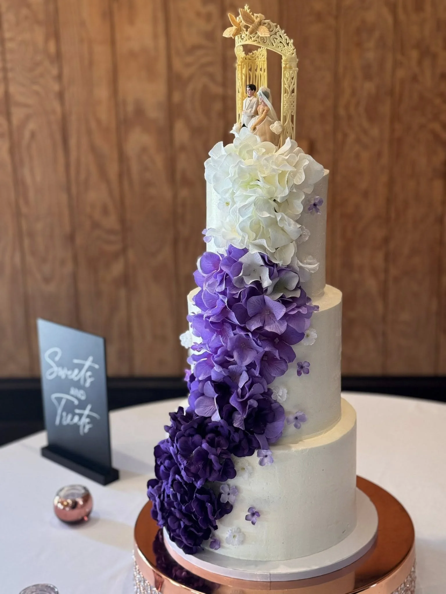 A wedding cake decorated with white and purple hydrangeas, topped with a figurine of a bride and groom under an ornate gold arch.